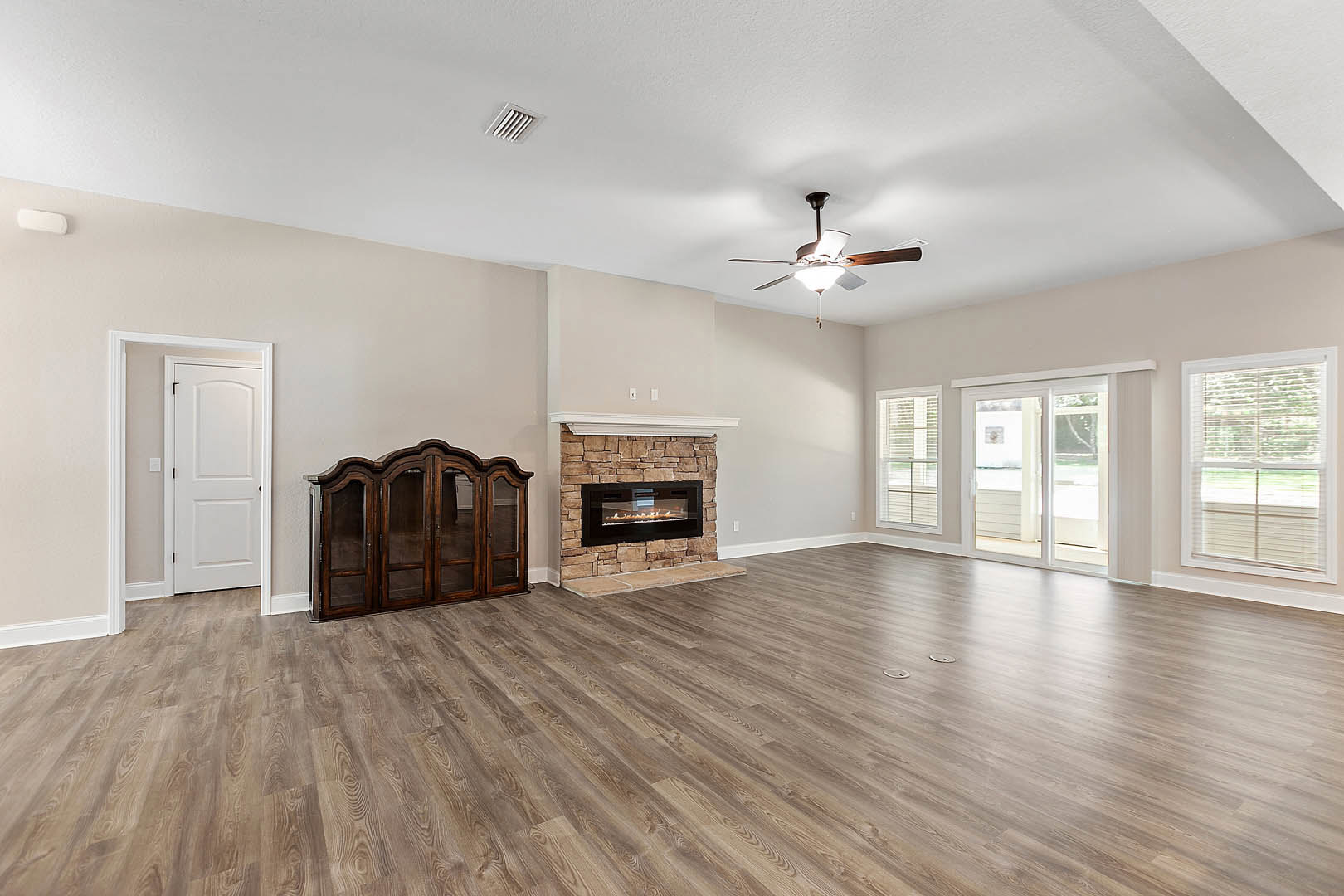 Living room with wood flooring, stone fireplace with burning fire, ceiling fan with light, wooden cabinet with glass doors, window with white blinds, and white door with black