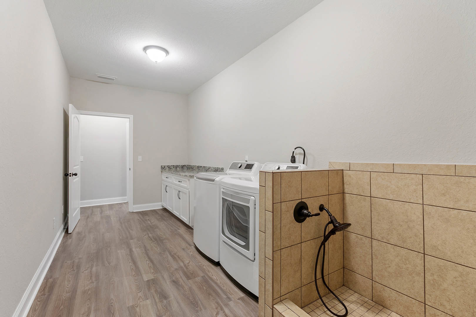 Laundry room featuring wood plank flooring, white tile walls, built-in cabinetry, a white door with black handle, ceiling light fixture, and a wall-mounted shower head with hose.