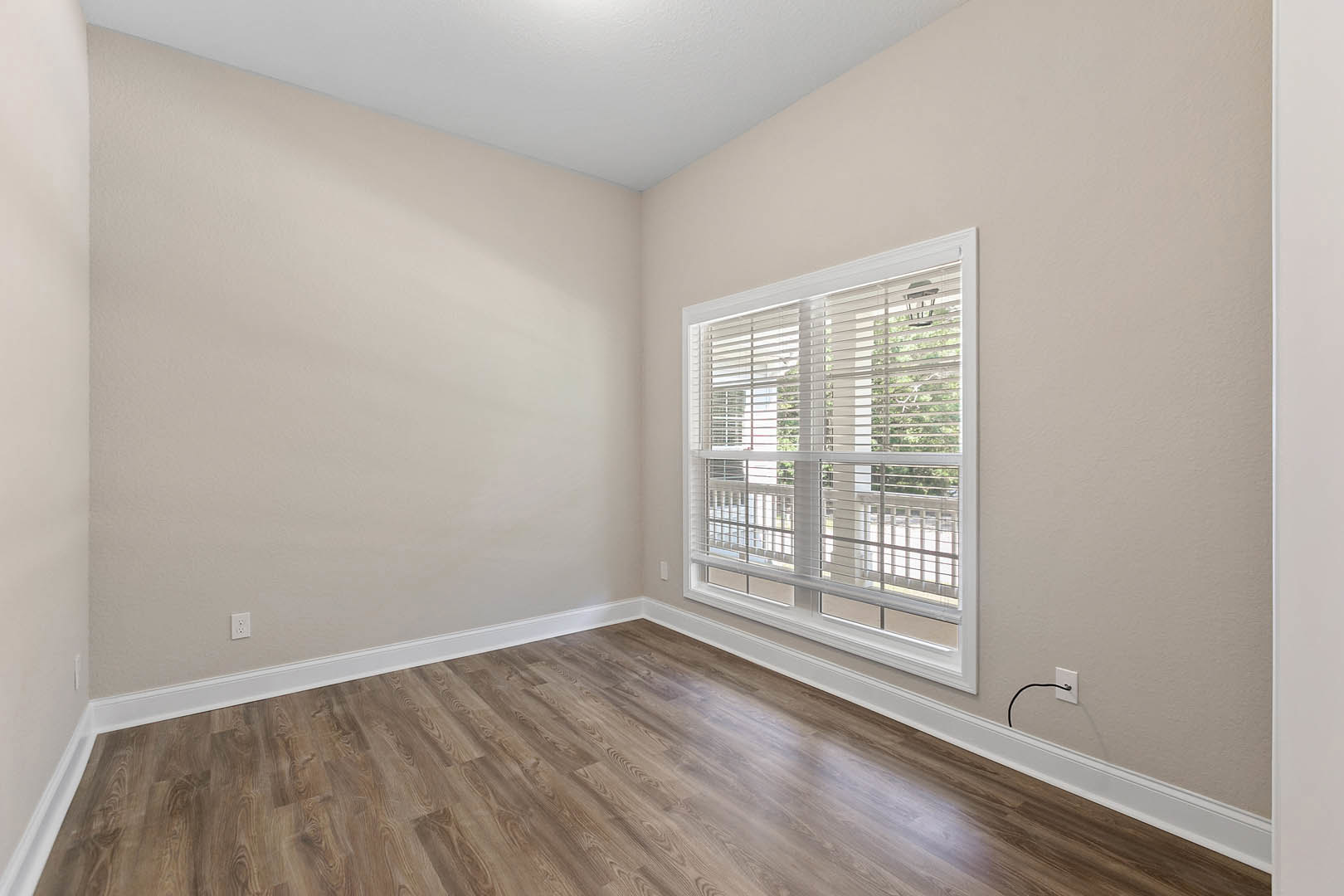Sunlit room featuring a large window with white blinds, smooth white walls and ceiling, and polished hardwood flooring with white baseboard trim