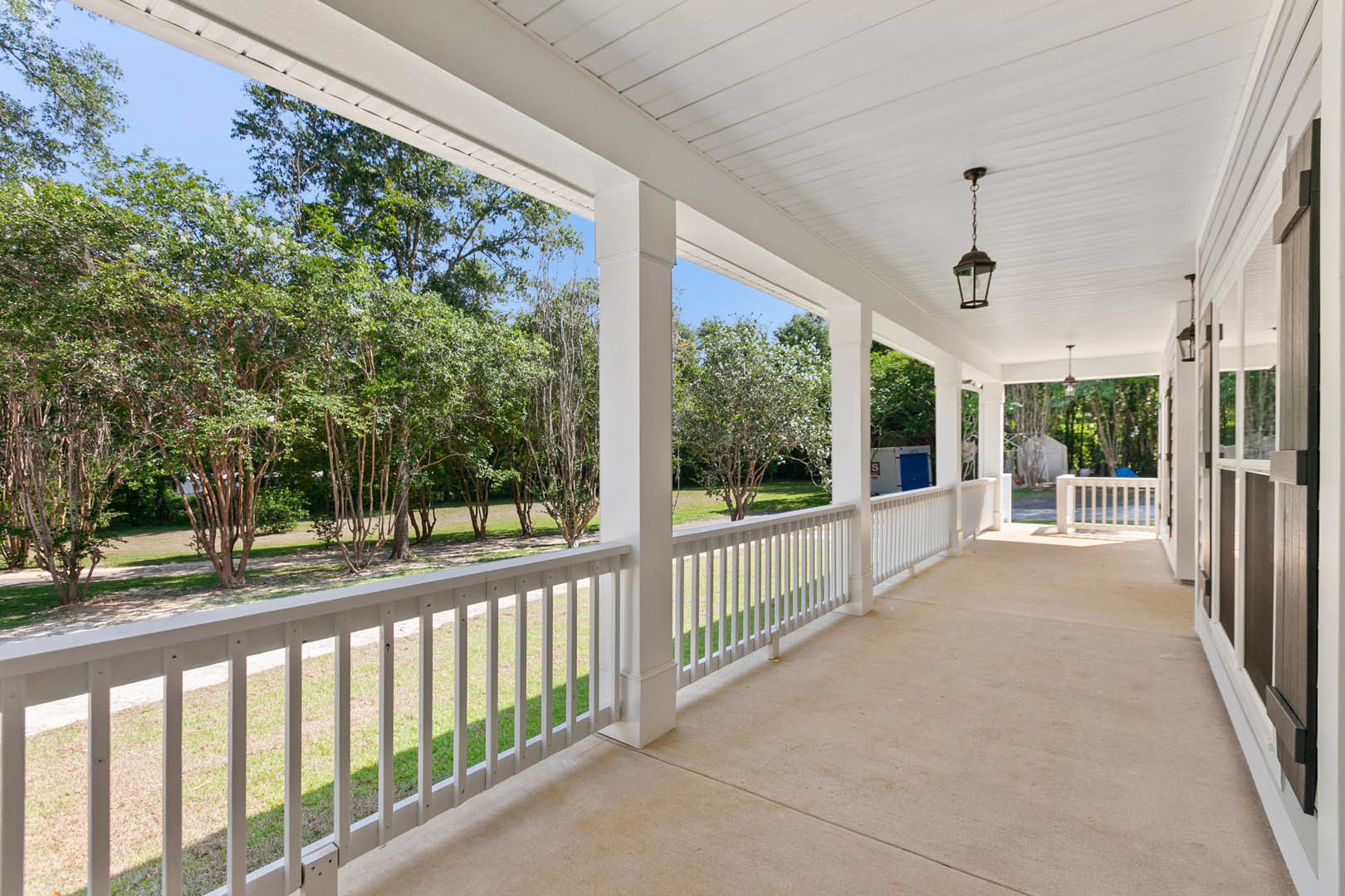 White porch with pillars and baluster railing overlooking a green lawn, surrounded by trees under a clear sky