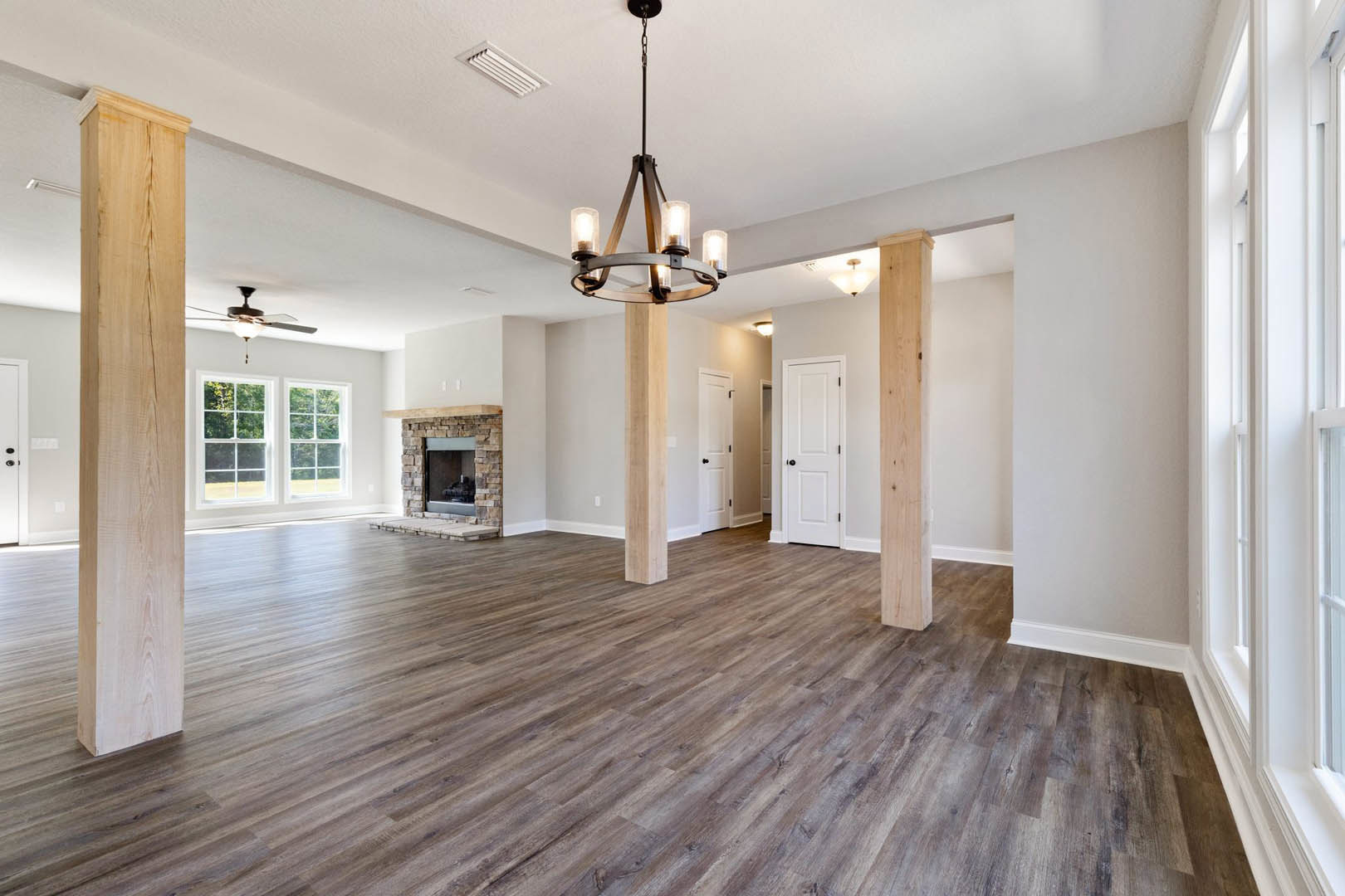 Spacious room with light wood flooring, white walls, central fireplace with stone surround, white door featuring black hardware, large window, modern chandelier, and detailed wood