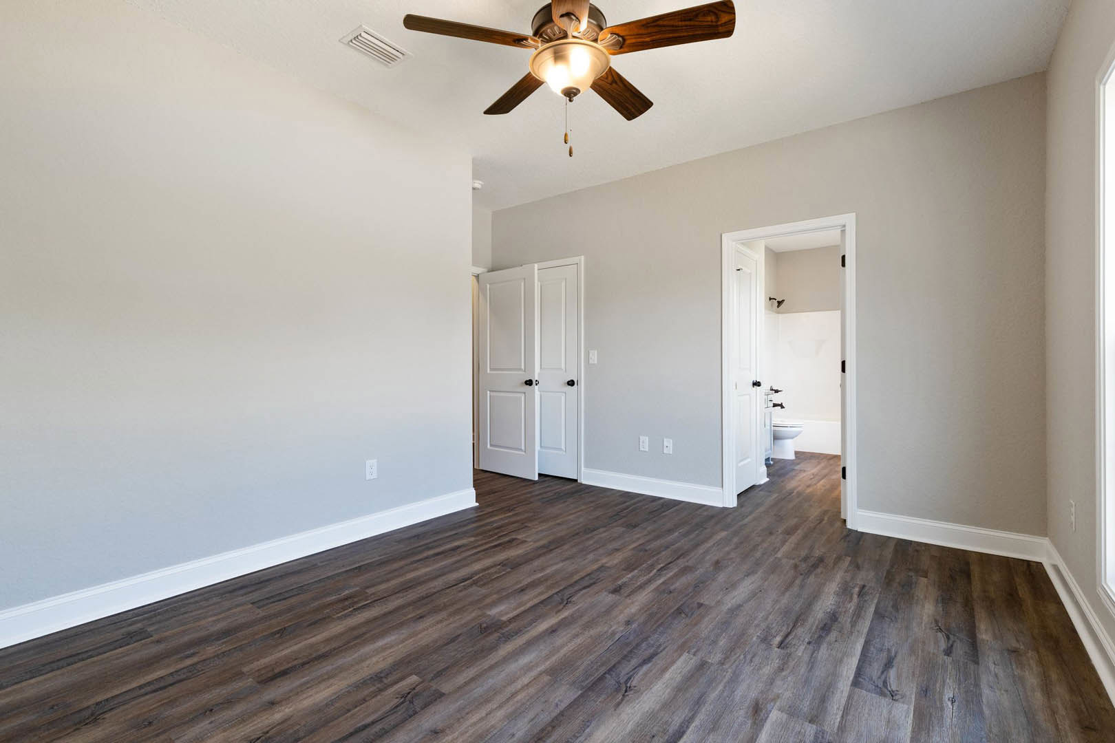 Ceiling fan with light fixture mounted above wood flooring, white walls, white door with black knobs, and adjacent white bathroom featuring toilet and tub