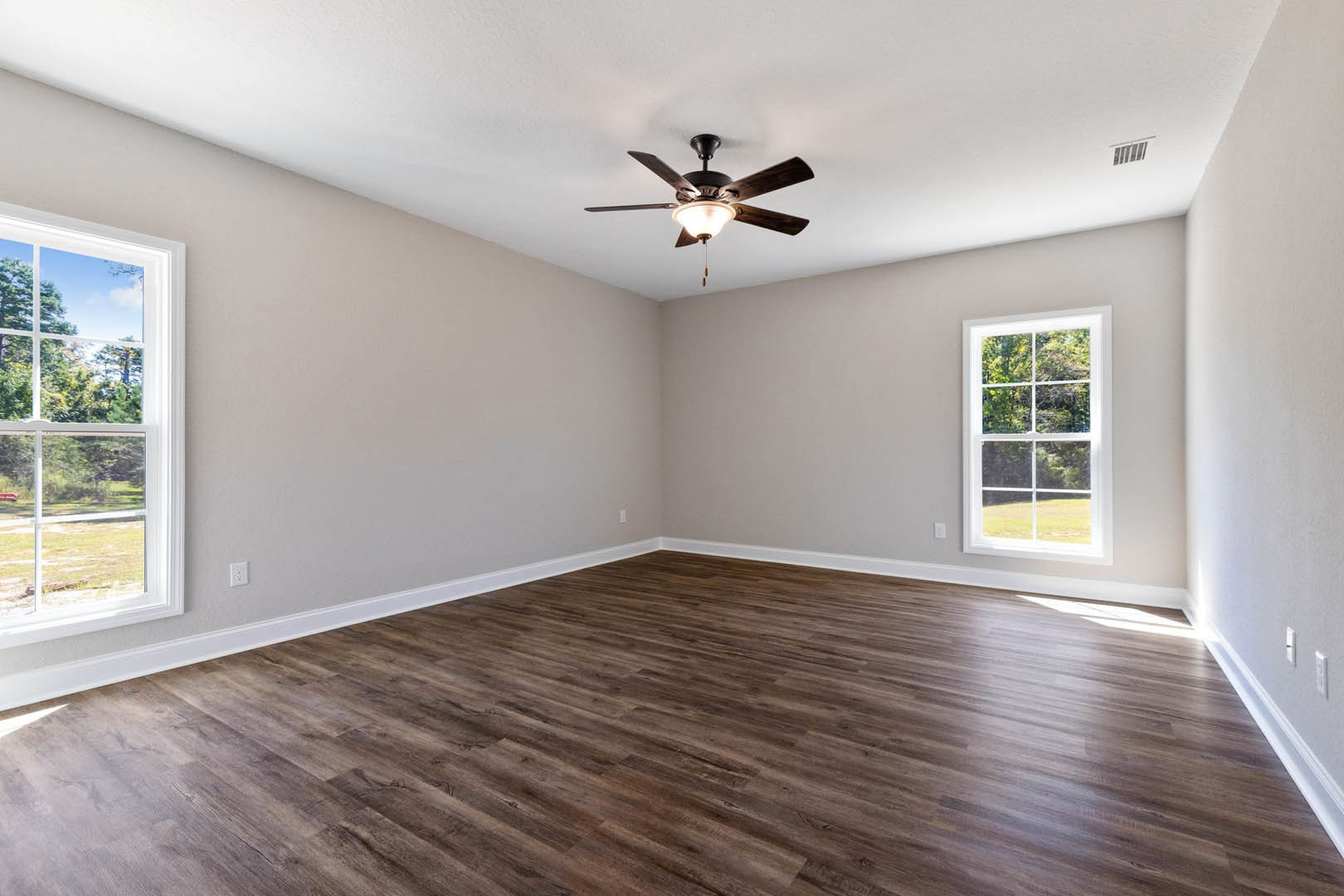 Ceiling fan with light fixture above hardwood floor, white baseboard trim, large window showing leafy trees outside