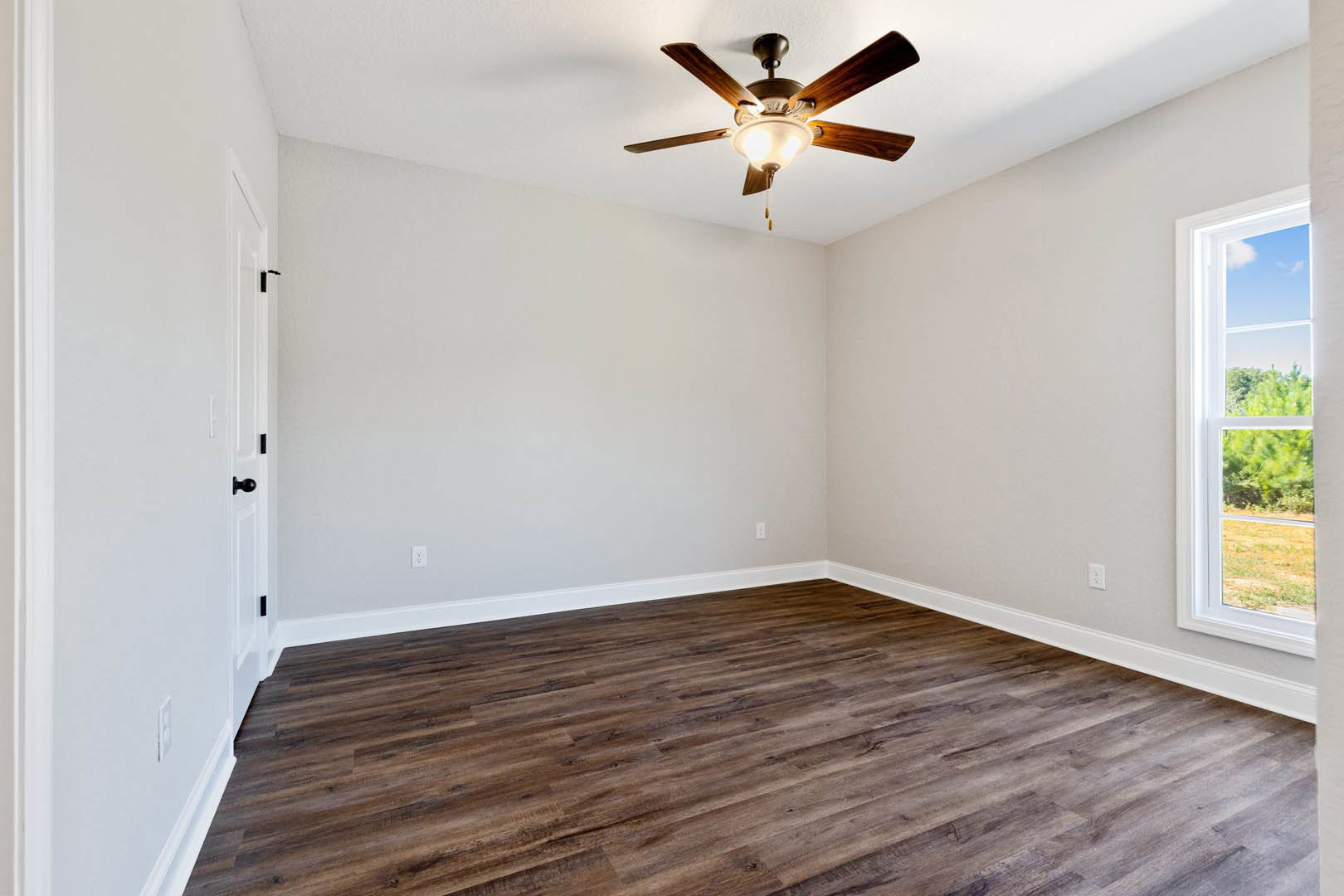 Ceiling fan with light fixture centered in a room featuring hardwood floors, neutral walls, and a window overlooking trees.