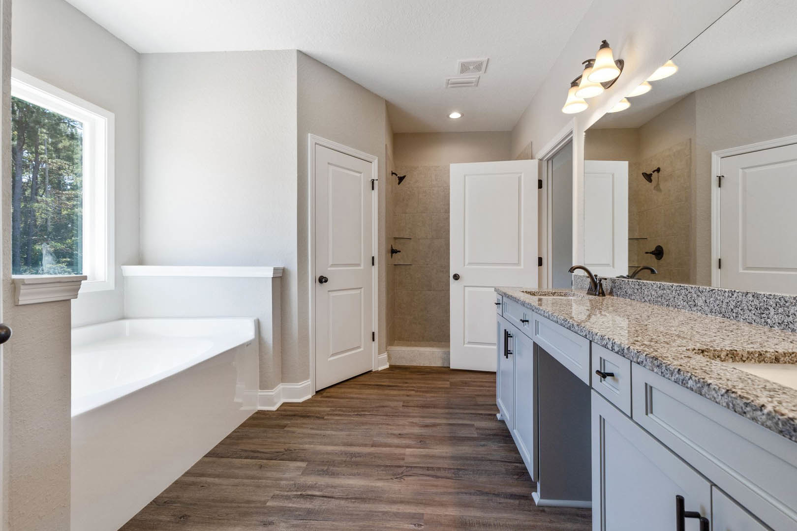 Bathroom with freestanding bathtub, white vanity and sink, wood flooring, white door with black hardware, window overlooking trees, light tile walls