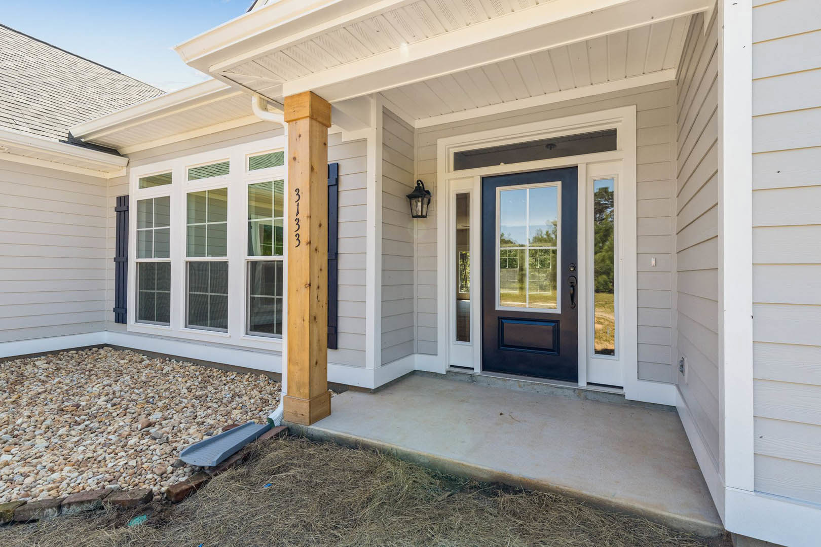 Blue front door with glass panels, stone walkway bordered by gravel, wooden post displaying house number, porch lamp, siding, and large window reflecting trees.