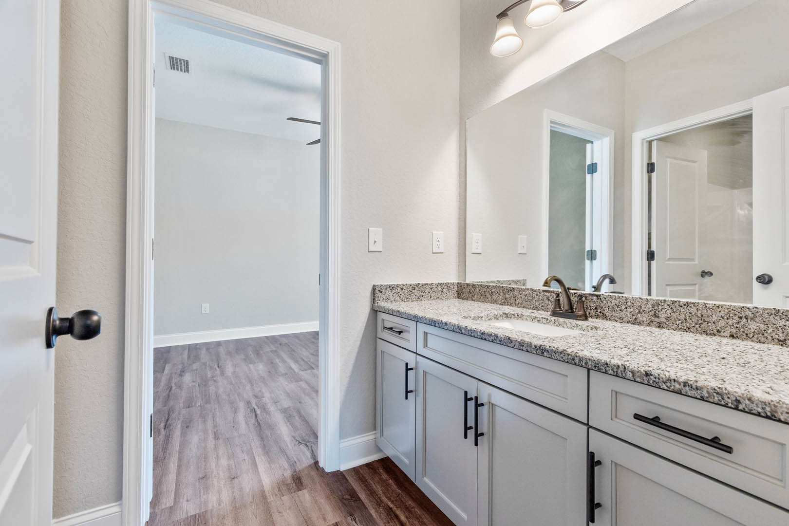 Bathroom with marble countertop, white cabinetry featuring black handles, wood flooring, open white door, modern sink, and close-up of a light fixture.