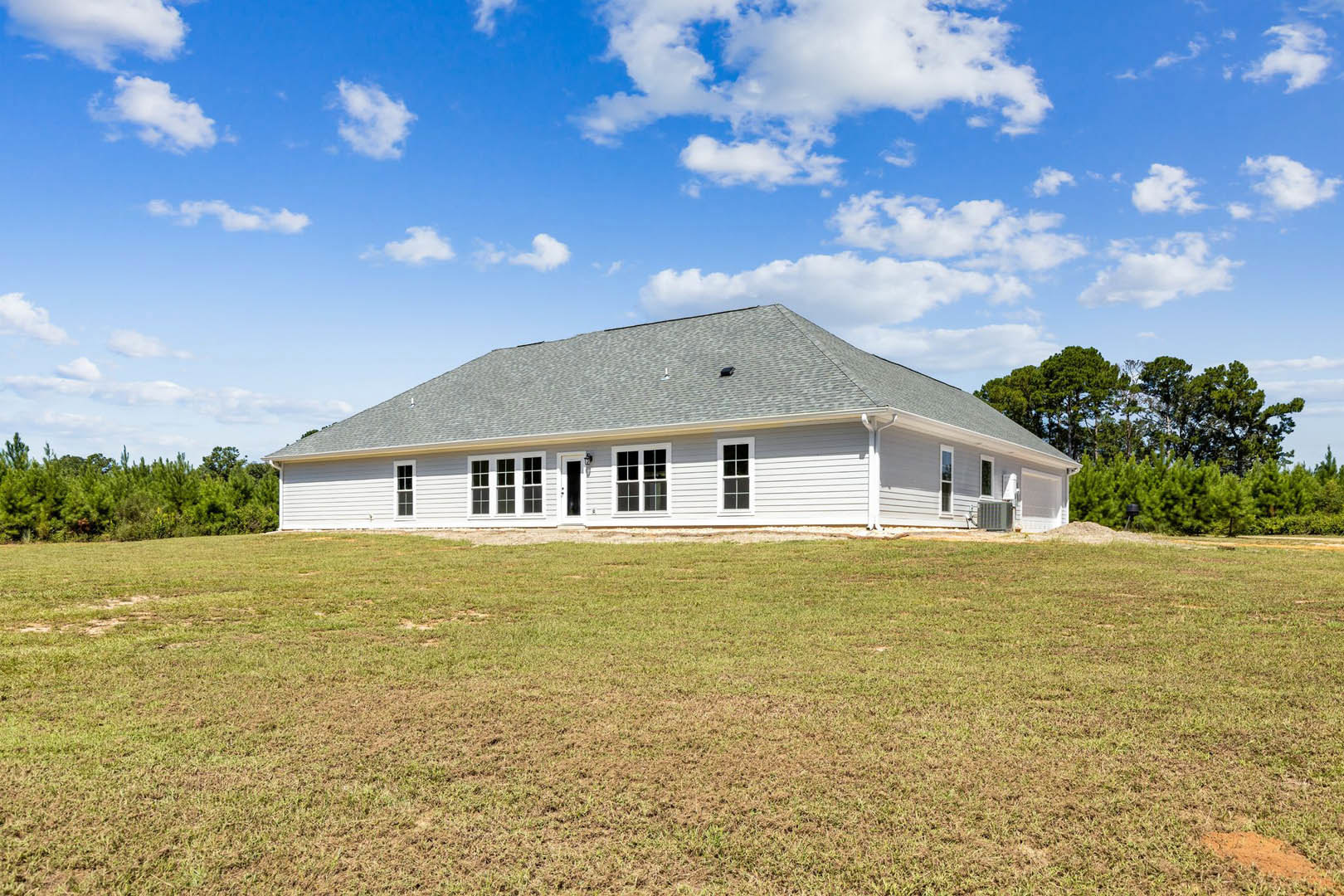 White farmhouse with gabled roof, large green lawn, blue sky with scattered clouds, mature trees bordering property