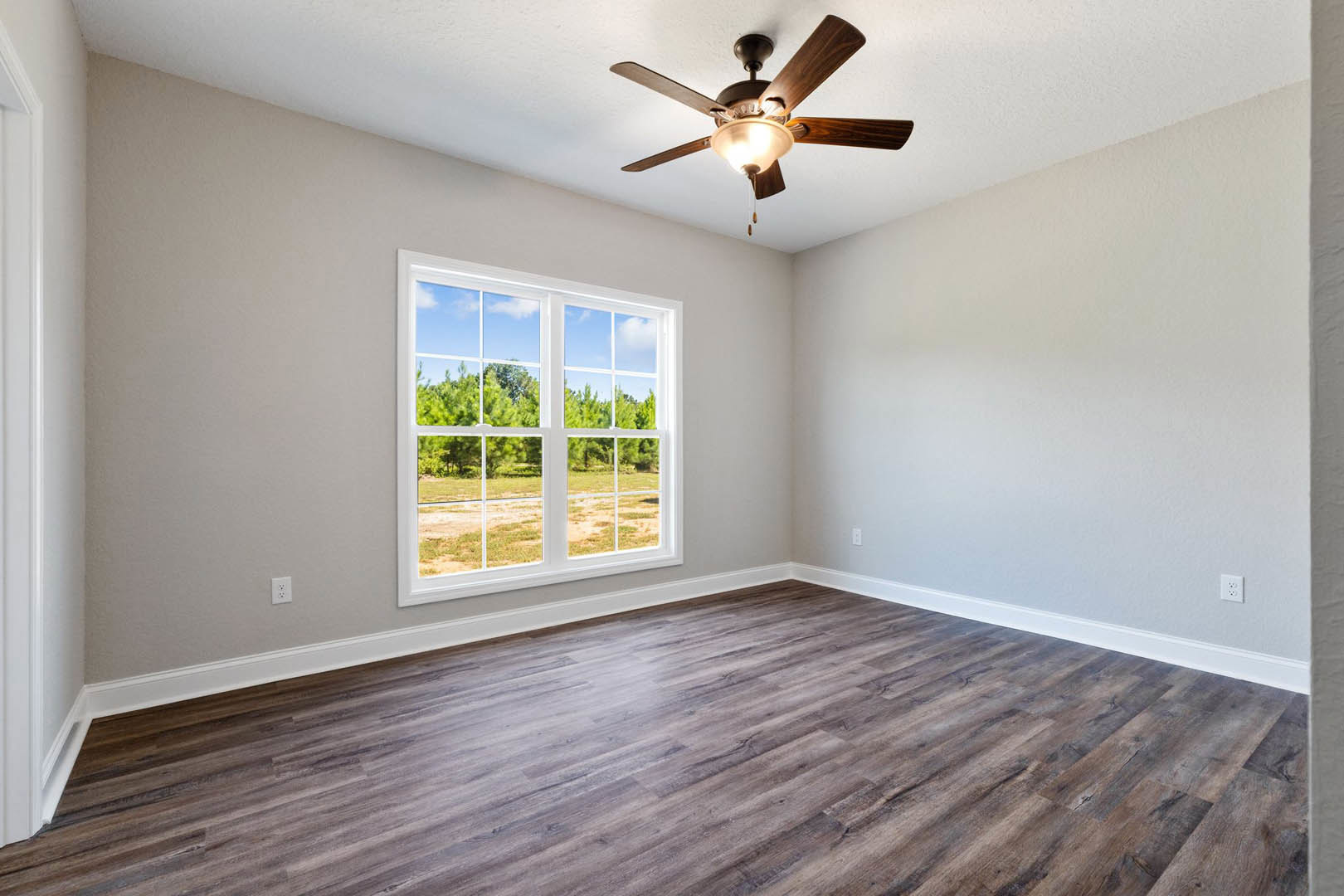 Ceiling fan with light fixture above wood flooring, large window with view of trees, neutral walls and trim