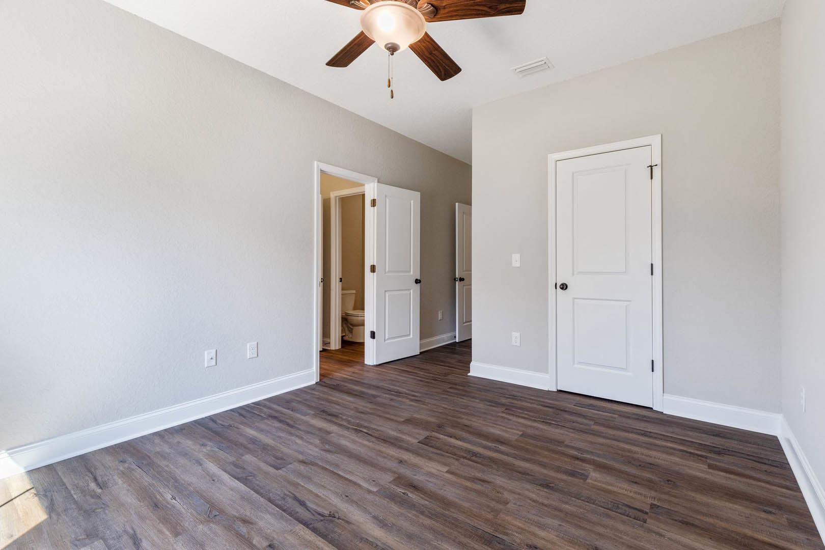 Wood flooring and white walls in a room with a ceiling fan and light fixture, white door with black handle, and simple modern finishes