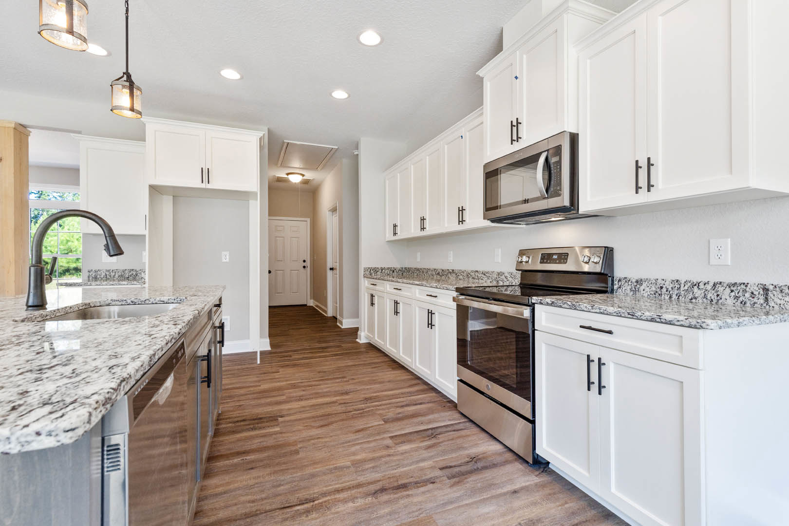 White kitchen cabinets, granite countertops, stainless steel stove and oven, open microwave door, chrome faucet, white electrical outlet on backsplash, light-colored flooring