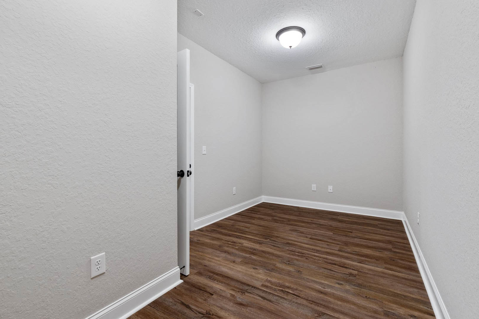 Room with light wood flooring, white walls, white door, ceiling light fixture, and electrical outlet visible near the baseboard.