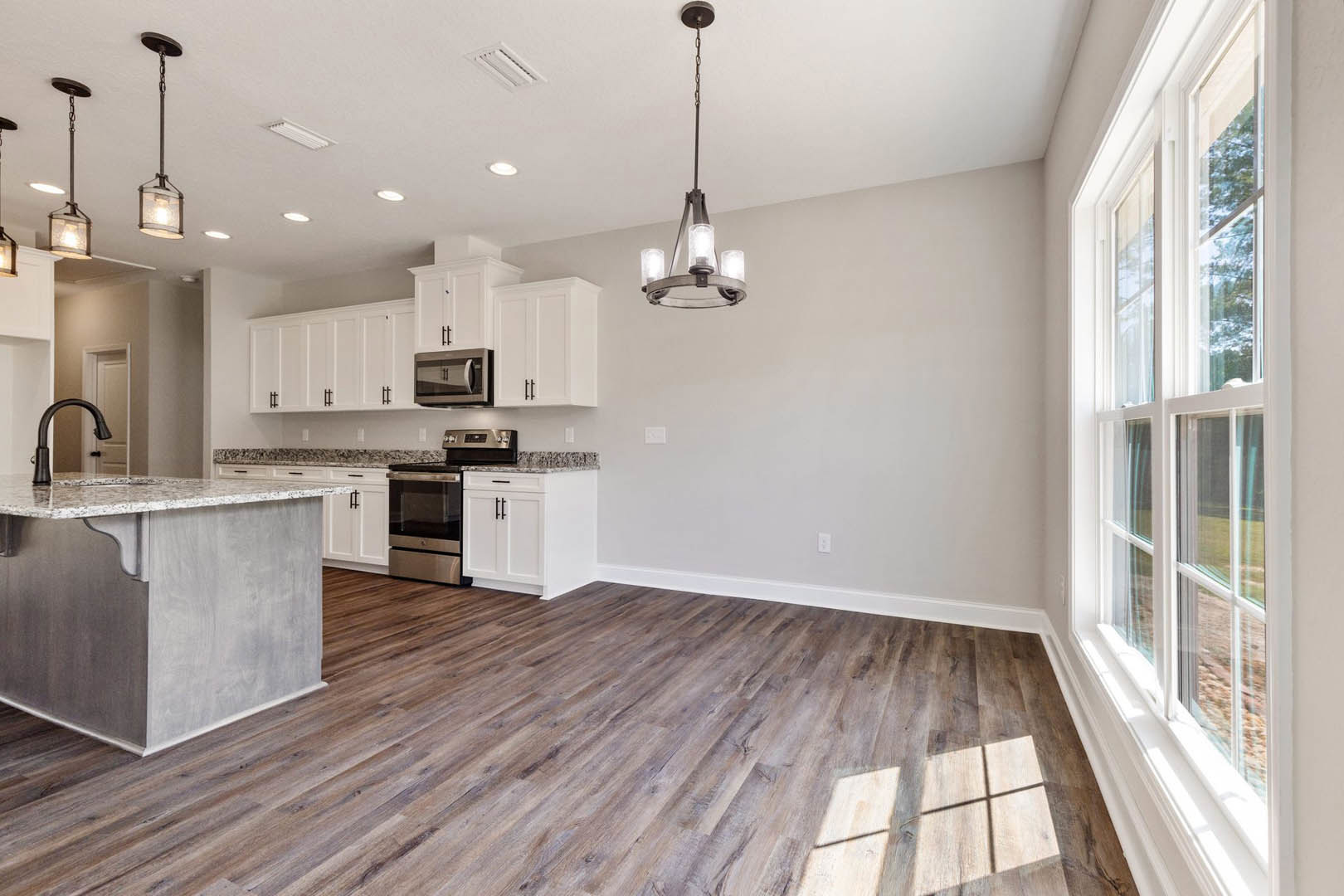 Kitchen with wood flooring, large window with white frame, marble-topped island, stainless steel microwave, cabinetry, and metal light fixture.