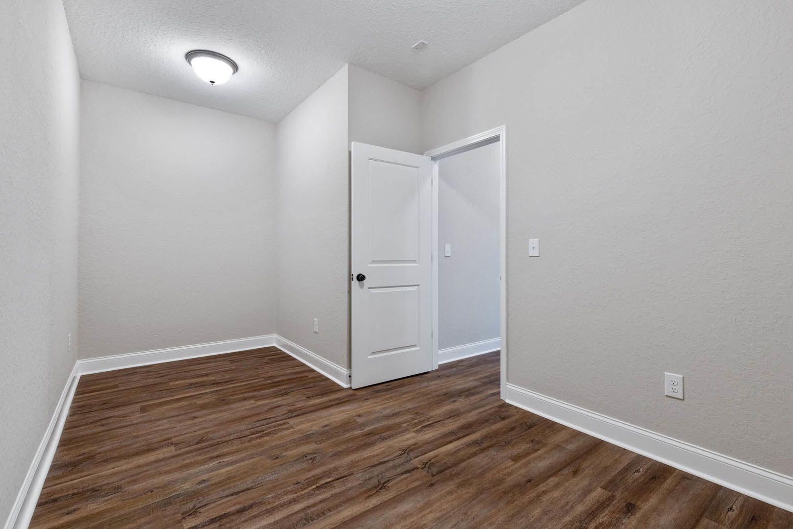 White paneled door with black knob open onto room with wood laminate flooring, white walls, ceiling light fixture, and visible white outlet and light switch.