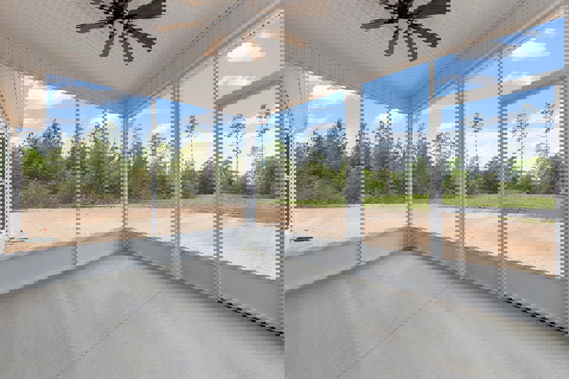 Open room with concrete floor, white pillars, ceiling fan with light, large windows overlooking dirt road and field of trees