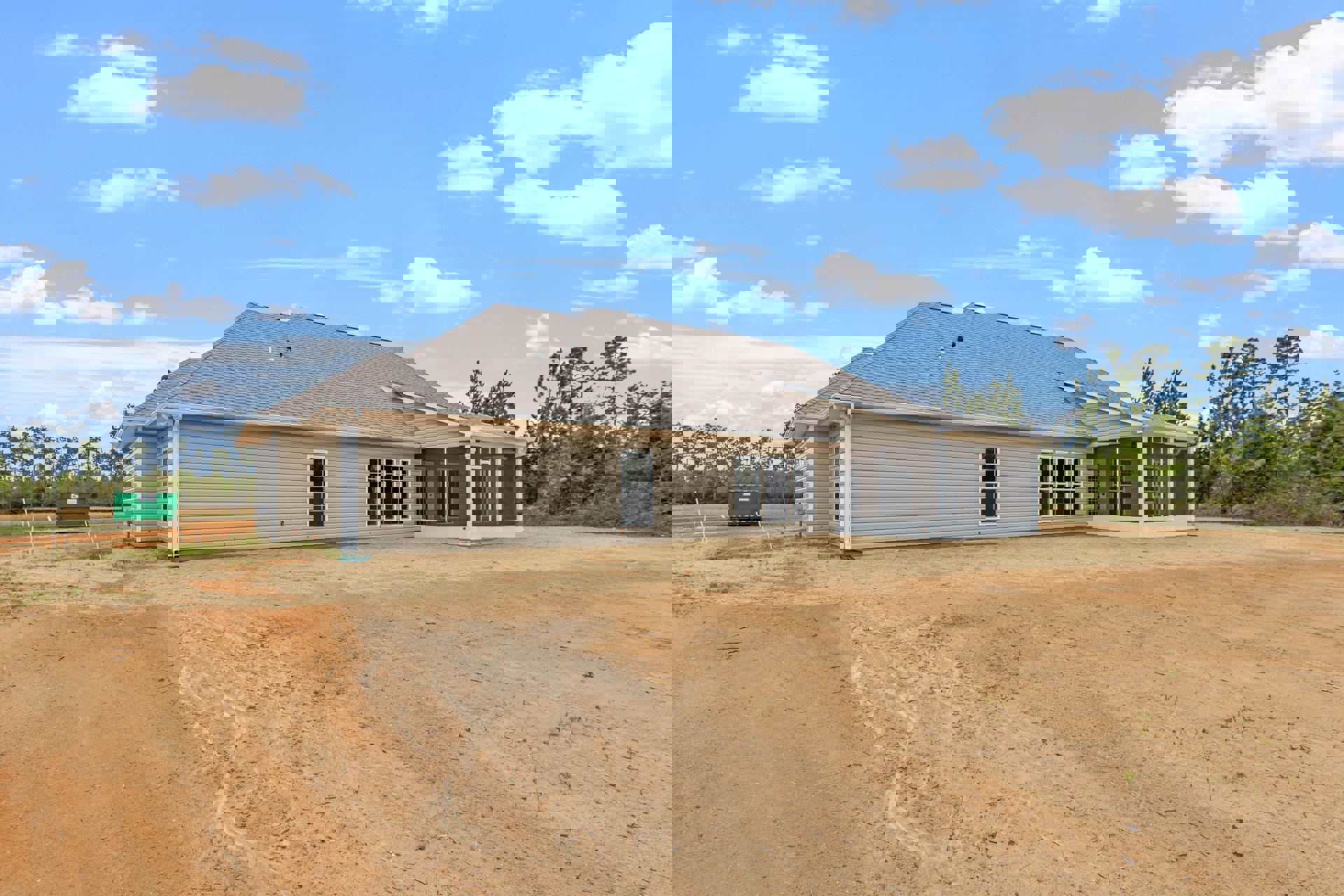 Two-story home with gray roof, row of windows, large dirt driveway, expansive grassy yard, green dumpster with white sign, mature trees, cloudy sky