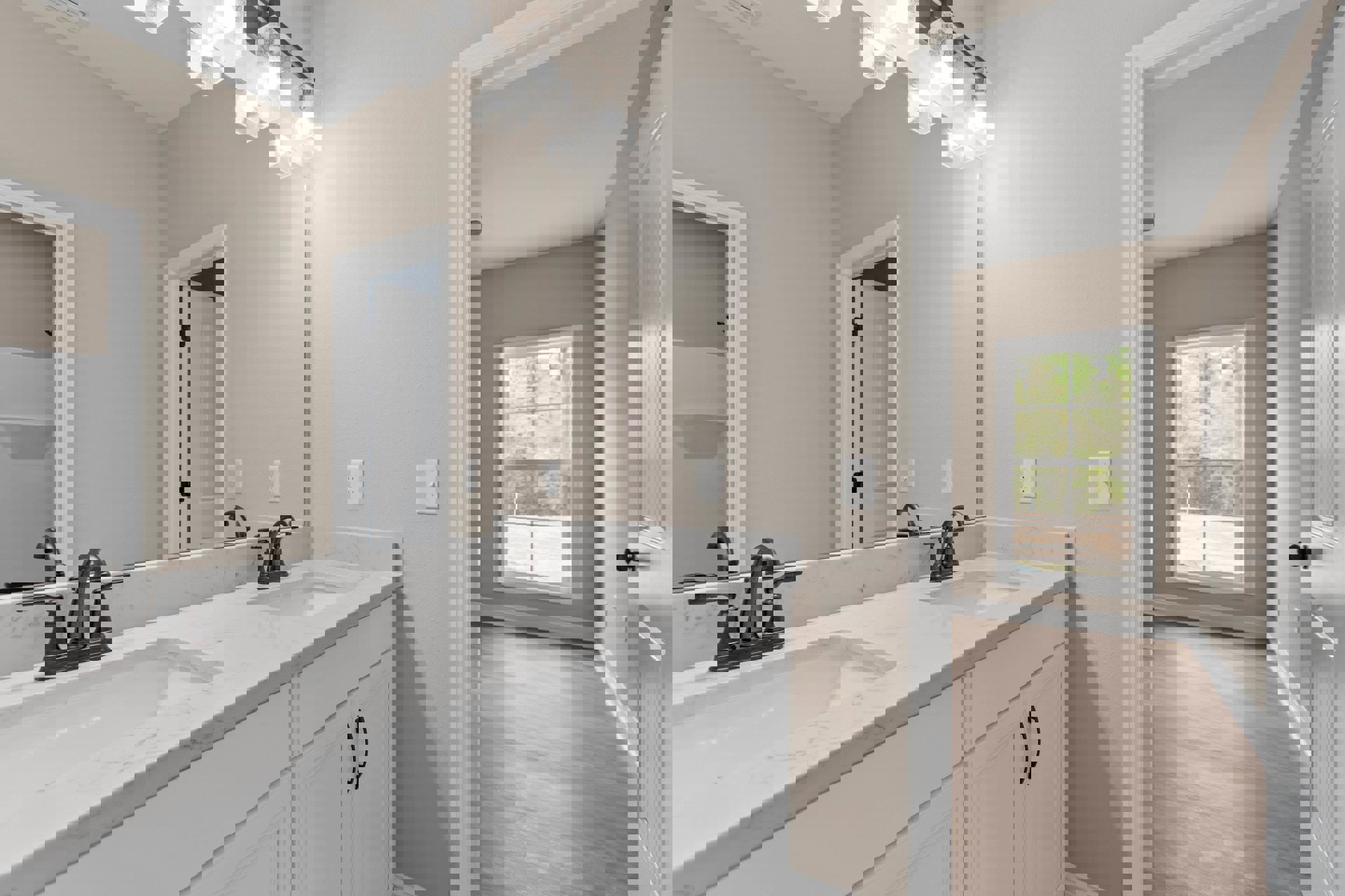 Bathroom with marble countertop, rectangular mirror above sink, chrome faucets, white walls, wood flooring, window showing trees outside, white door with black hinge, light switch