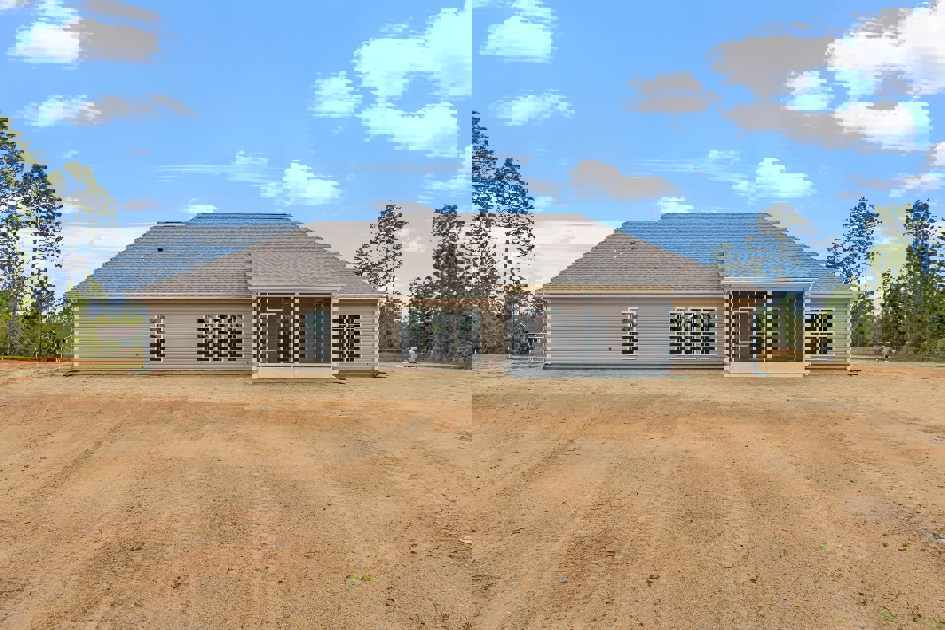 Single-story home with a large roof, row of windows, surrounded by a dirt field with visible tire tracks, under a blue sky with scattered clouds.