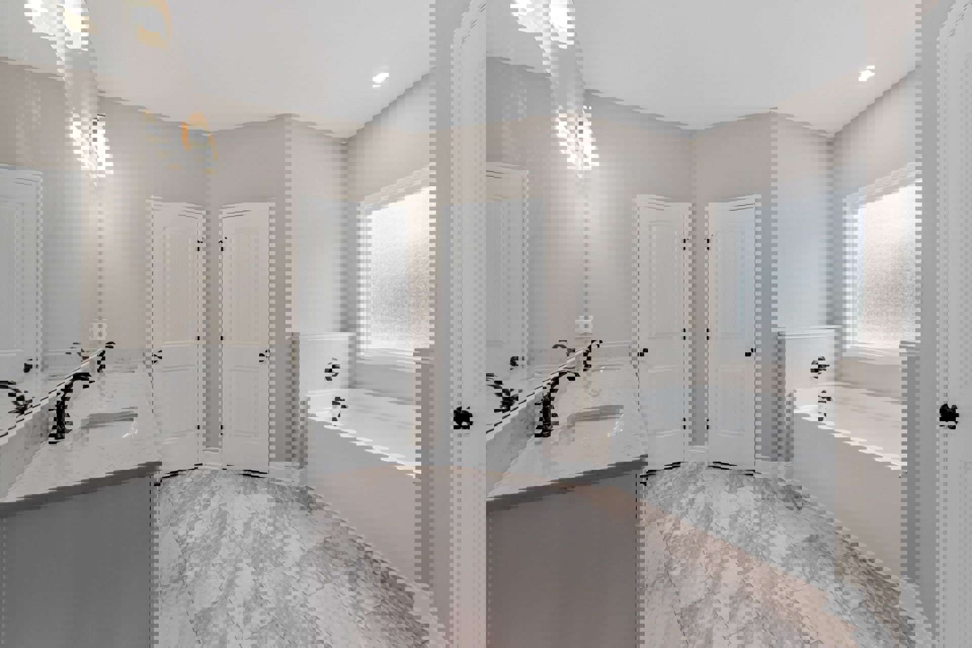 Marble tile flooring, white walls, white ceiling, freestanding bathtub, white sink with chrome faucets, white door with black knob, window with trim