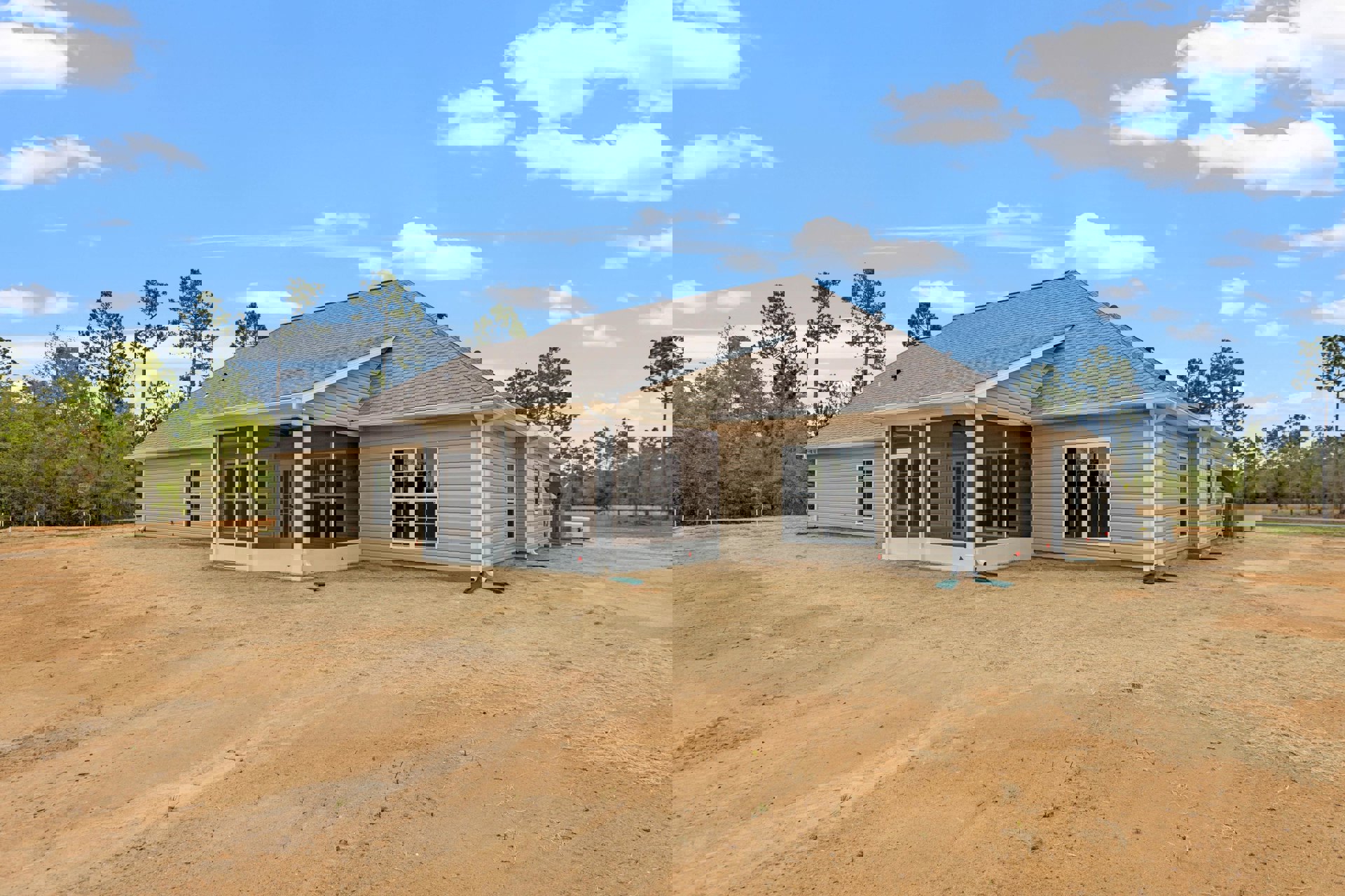Modern house exterior with gray siding, large windows, covered porch, and unfinished dirt yard surrounded by grass and trees under a cloudy sky