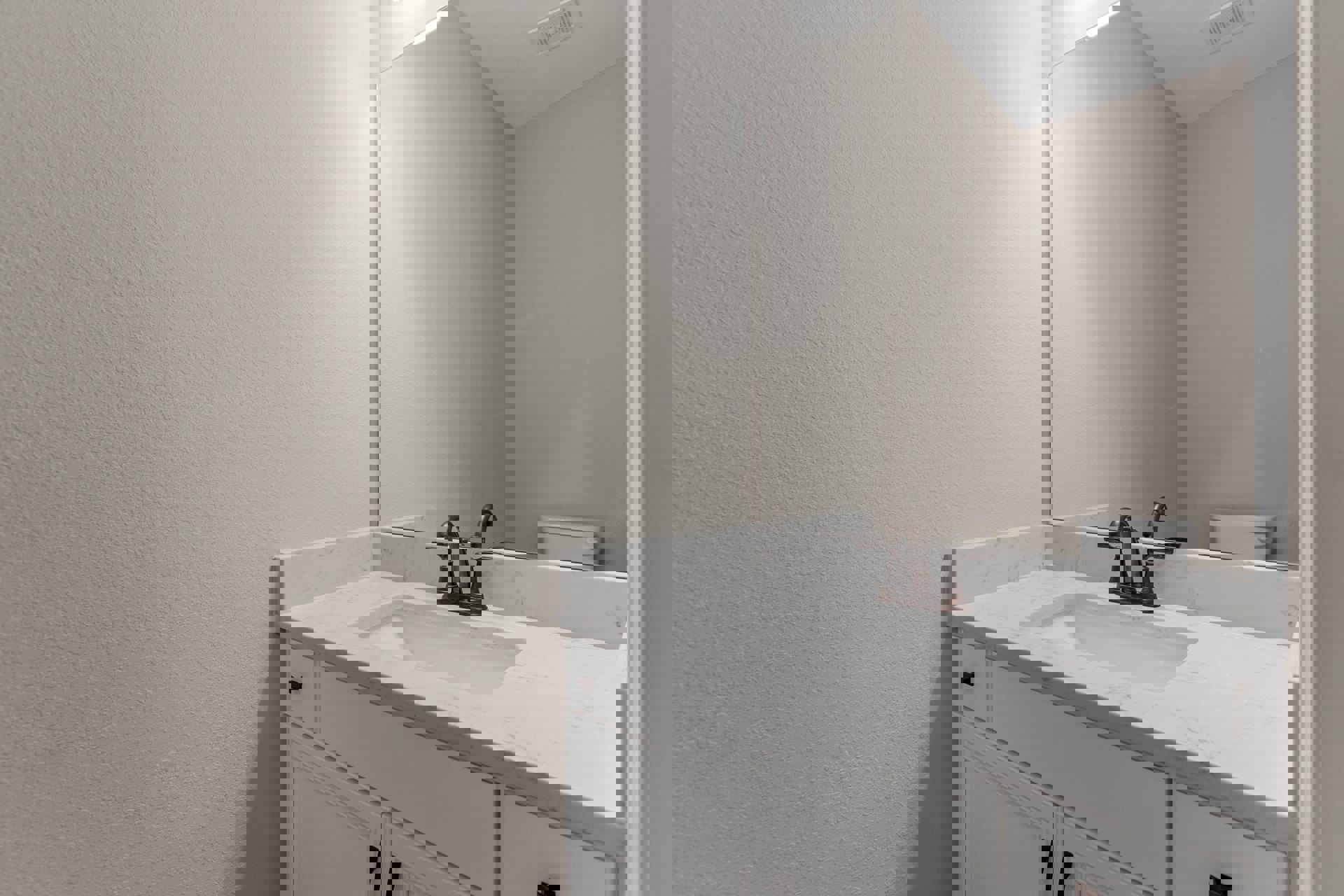 Modern bathroom with white walls, rectangular mirror above a sleek sink, chrome faucet, and visible ceiling vent