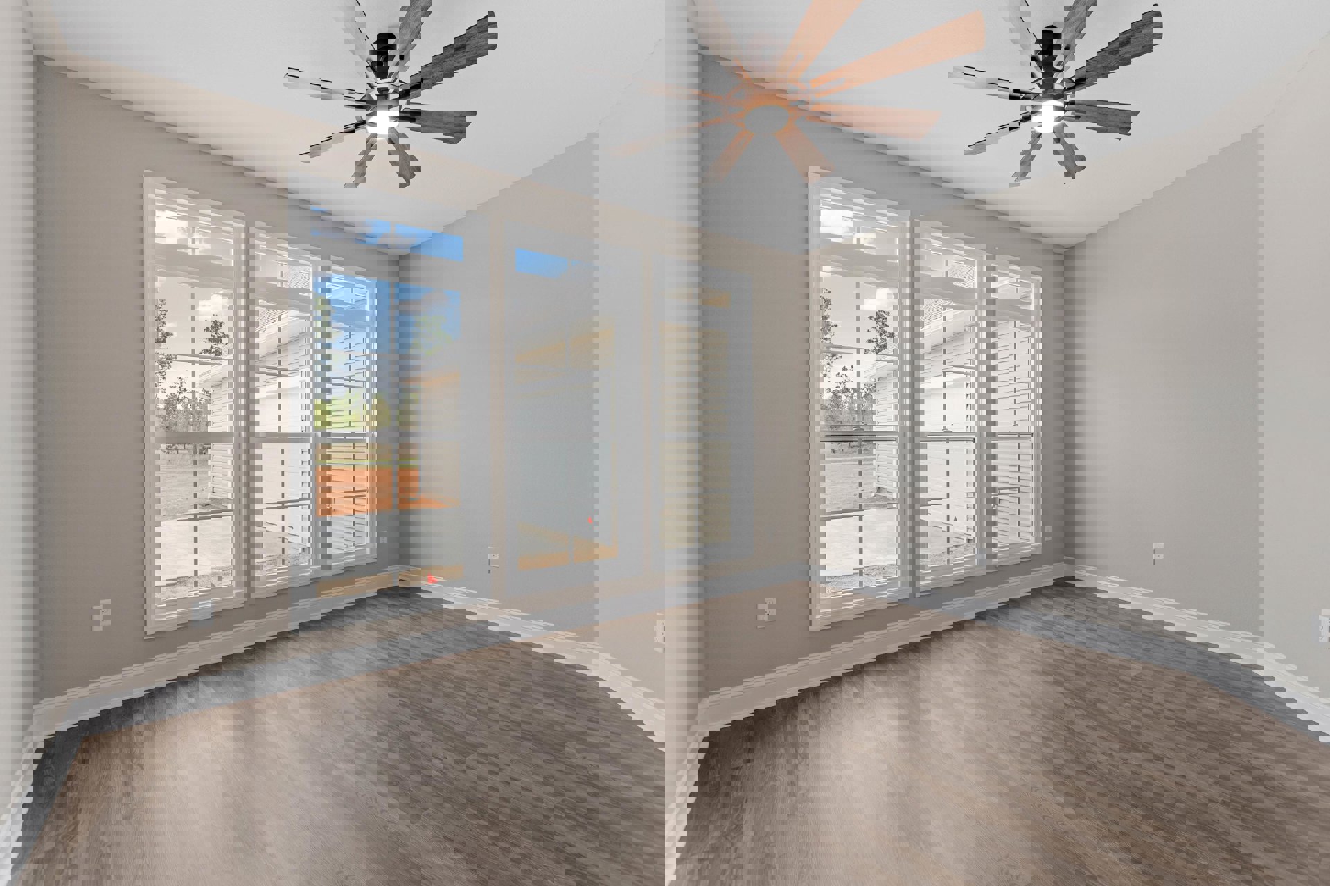 Bright room with wood flooring, white walls, ceiling fan with light fixture, large windows overlooking a dirt road and leafy trees