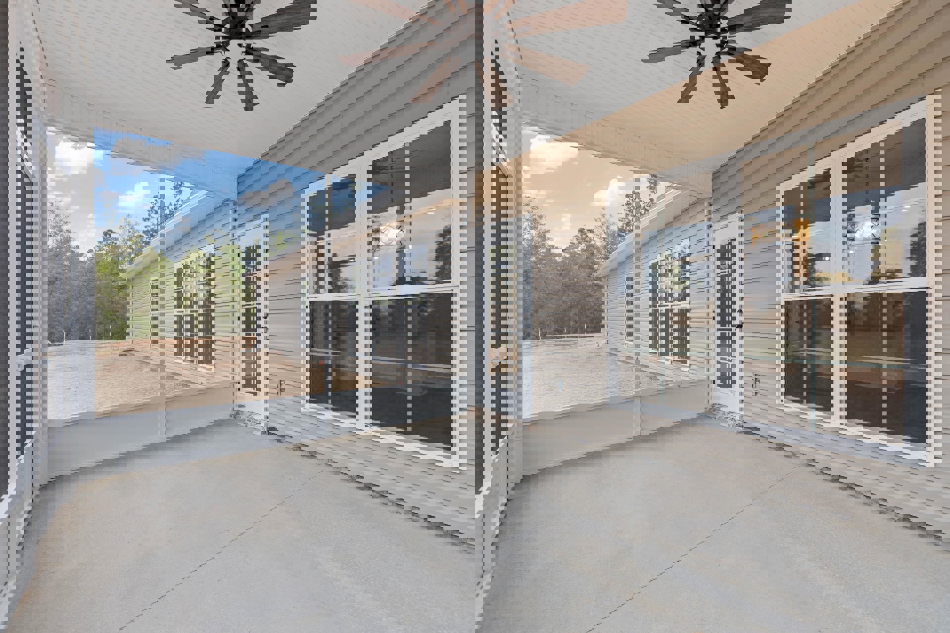 Covered front porch with white columns, ceiling fan with light, dark wood front door, garage door, concrete driveway, and large window on light-colored siding