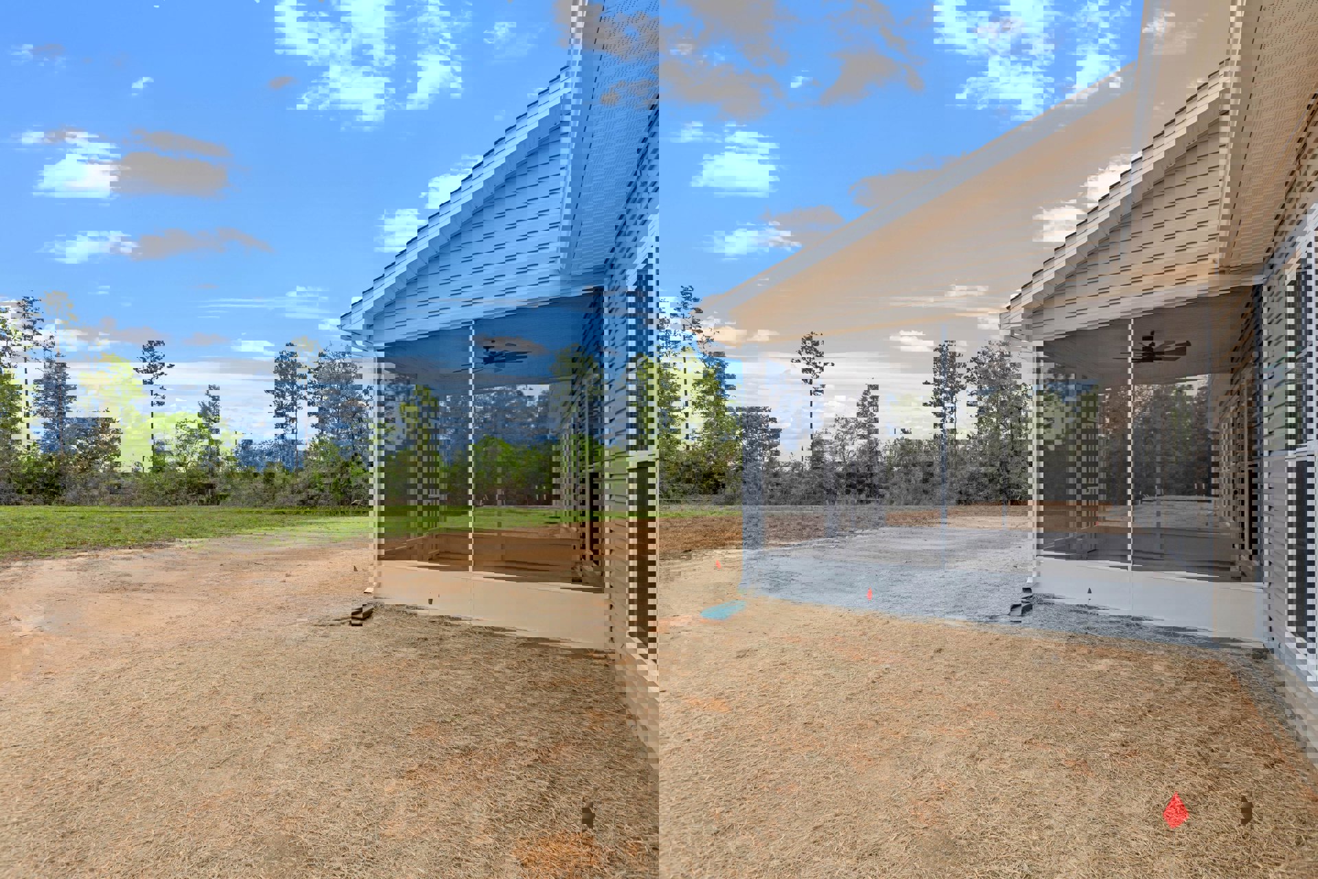 Screened porch with black ceiling fan, white trim, and screen door; green garden hose and red flag in grassy yard; trees and cloudy sky in background.
