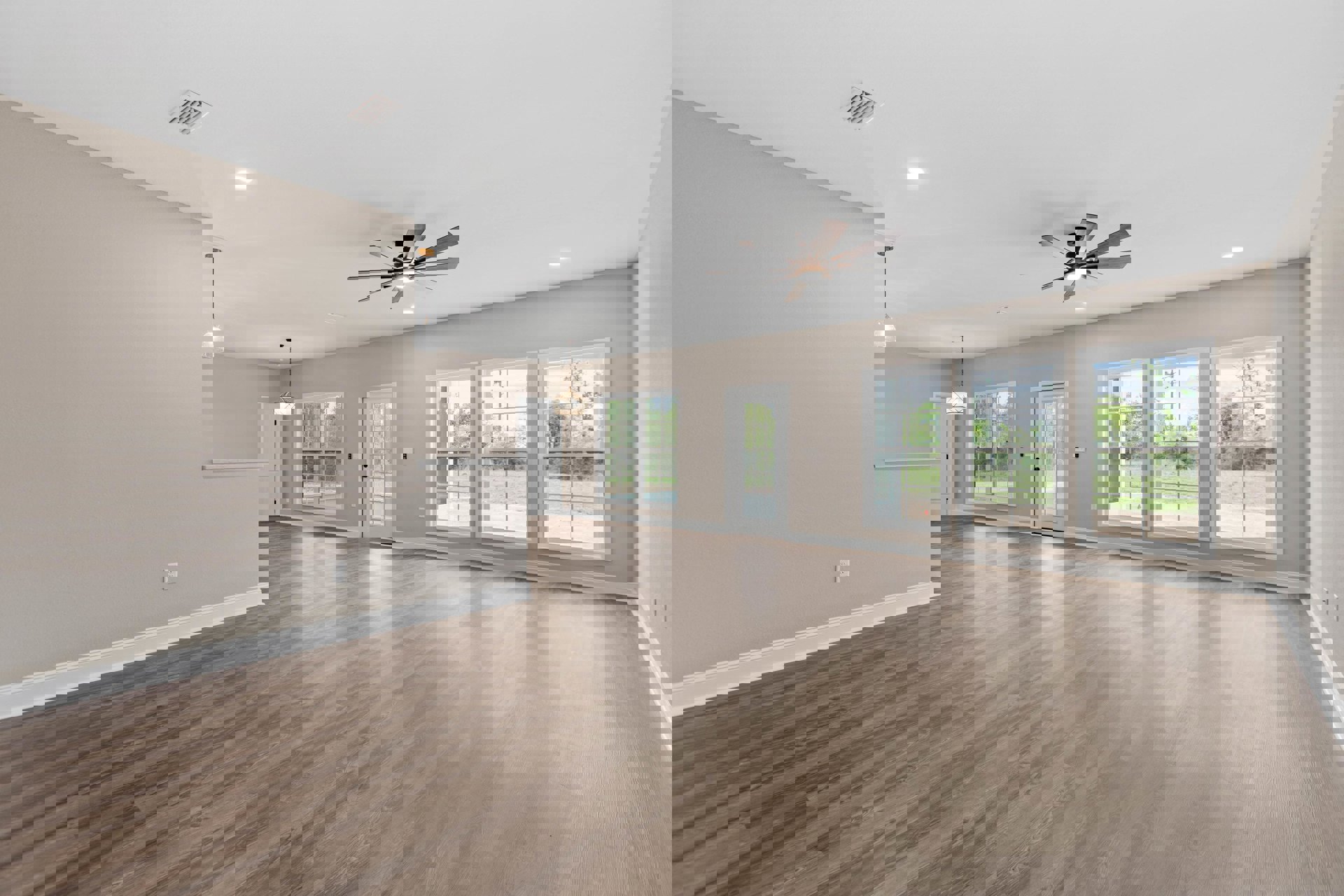 Spacious room with wood flooring, white walls, ceiling fan with light, multiple windows showing views of grass and trees, and a white door with glass panes