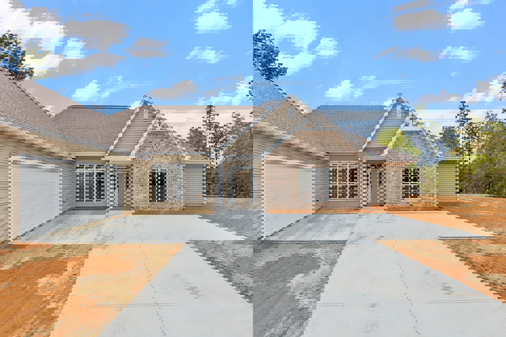 Brick exterior home with white-framed windows, tan garage door, paved driveway, and surrounding trees under a partly cloudy sky