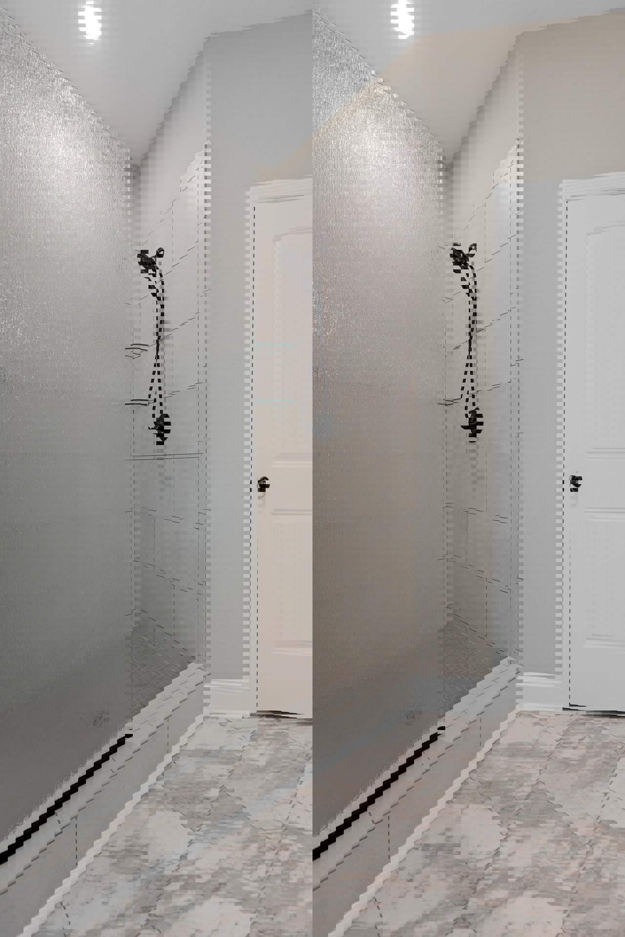 White tile shower with glass door, black shower head and pipe, built-in glass shelf, adjacent white door with black knob, ceiling light fixture, light-colored flooring