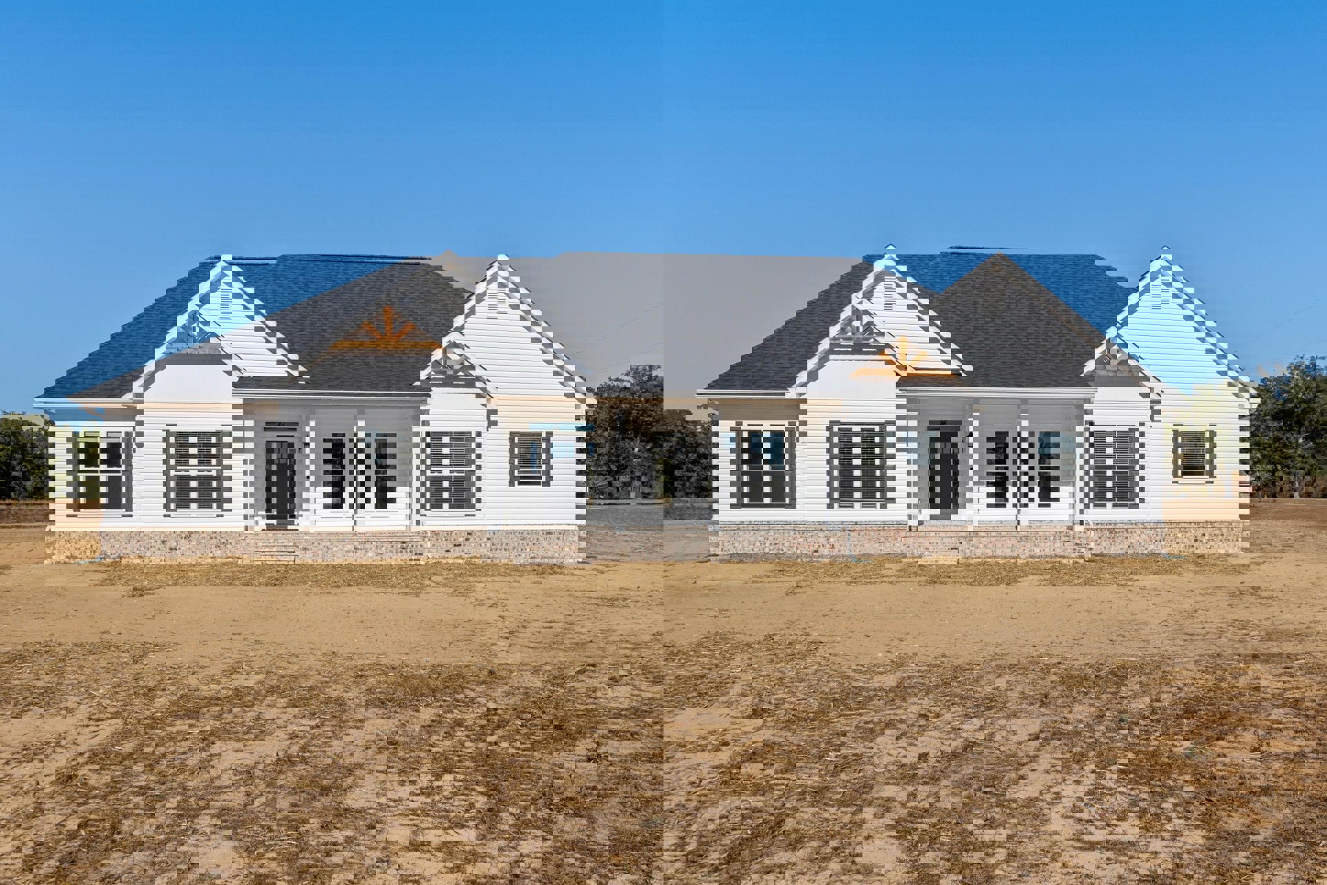 White house with columned front porch, black door with glass panel, window with closed blinds, surrounded by dirt field and sparse trees under clear sky