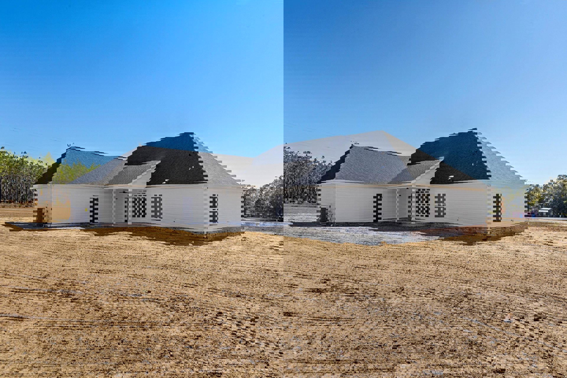 White house with covered porch, large dirt yard in foreground, grey HVAC unit beside exterior wall, blue sky overhead, window with closed blinds