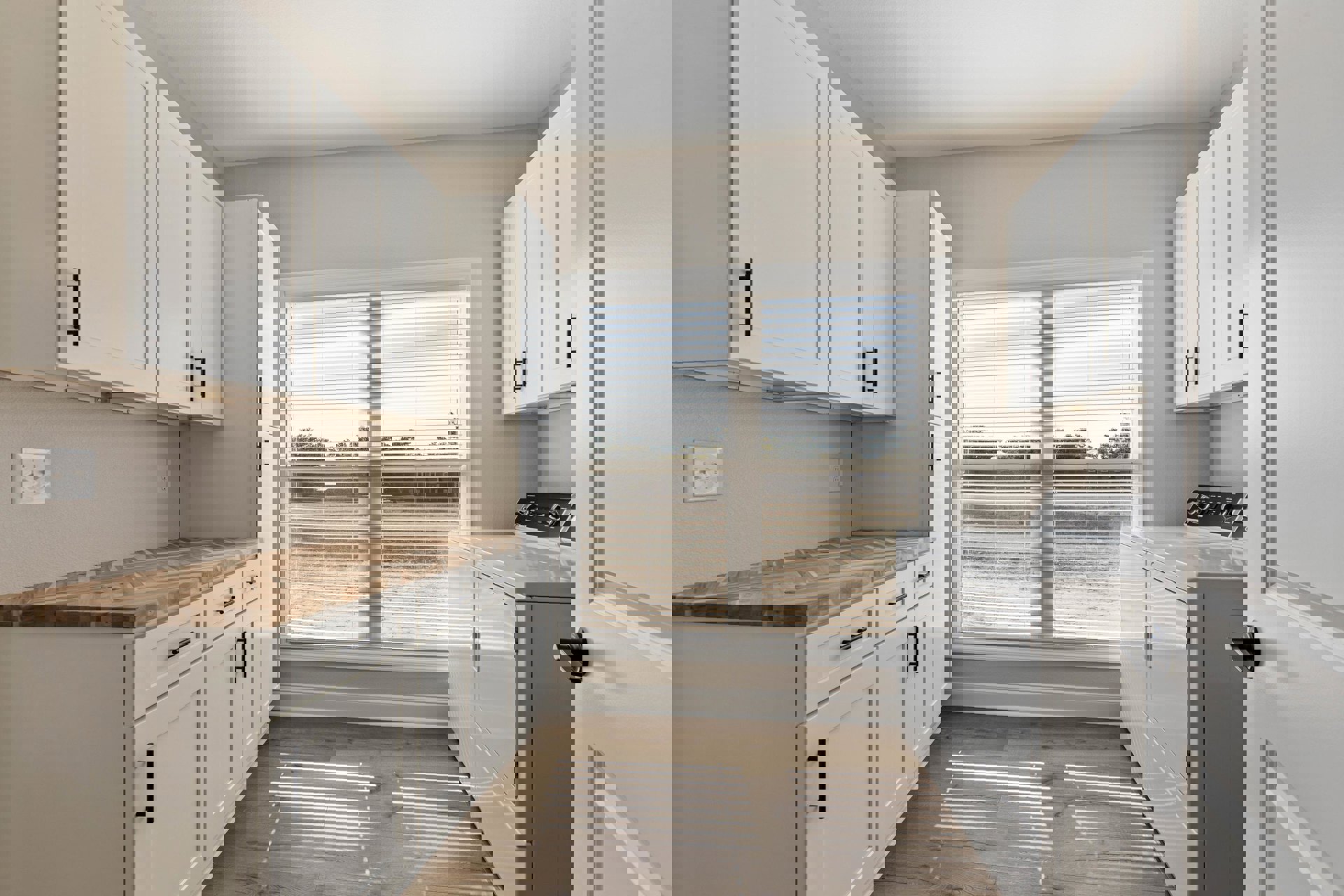 White cabinets with black handles, wooden floor, window with blinds, washing machine, countertop, and sink in a bright kitchen.