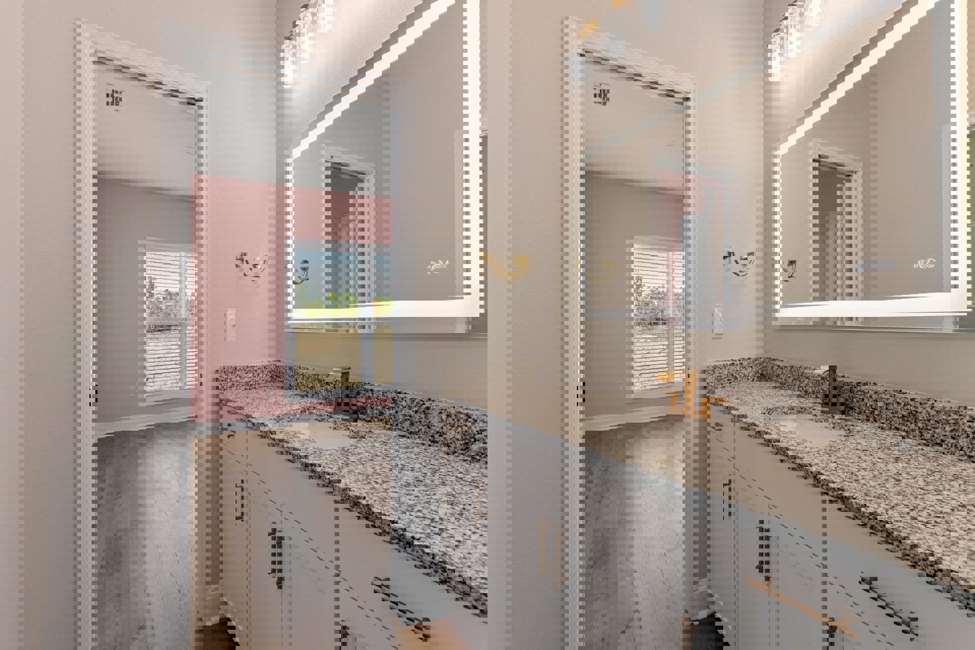 Bathroom with pink walls, marble countertop, undermount sink, white cabinetry, large mirror, window with blinds, chrome faucet, and modern light fixture.