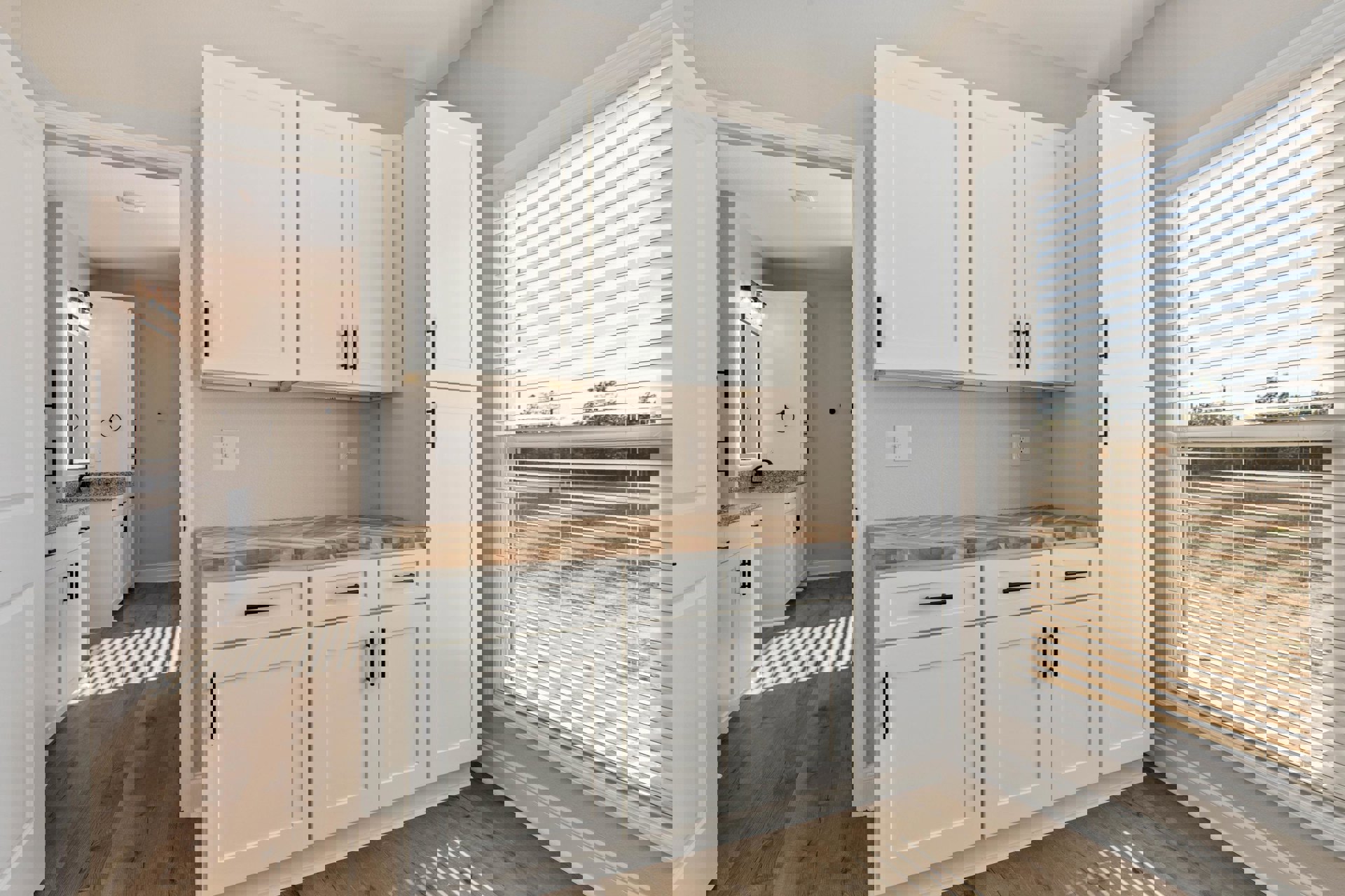 White kitchen cabinets with black handles, wood flooring, window with blinds, white door and wall, light switch on wall, countertops and drawers visible.