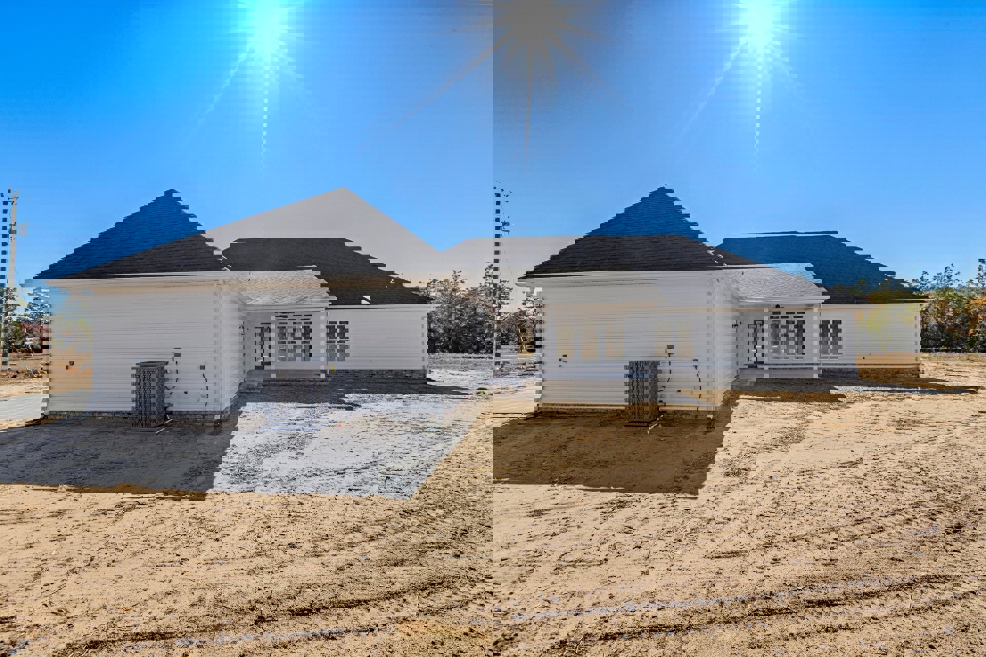 White house with black roof set on expansive green lawn, large grey metal box near driveway, telephone pole and leafy tree in background, bright sun overhead in clear blue sky