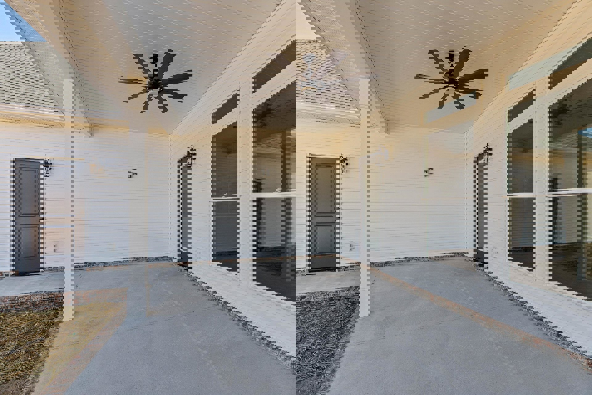 White exterior home with glass-paneled door, concrete patio, ceiling fan with light, brick edging, and grassy yard