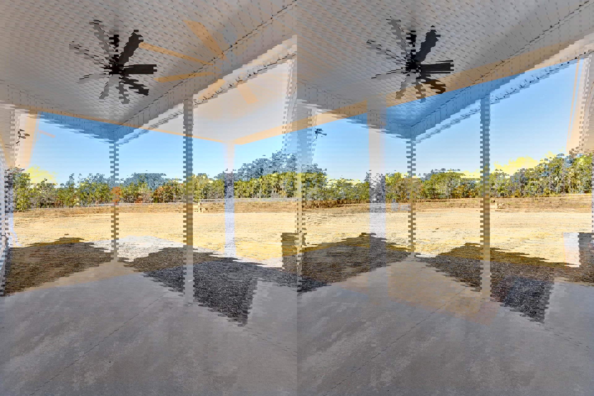 Covered patio with concrete floor, white pillars, ceiling fan with light, overlooking grassy field and trees.