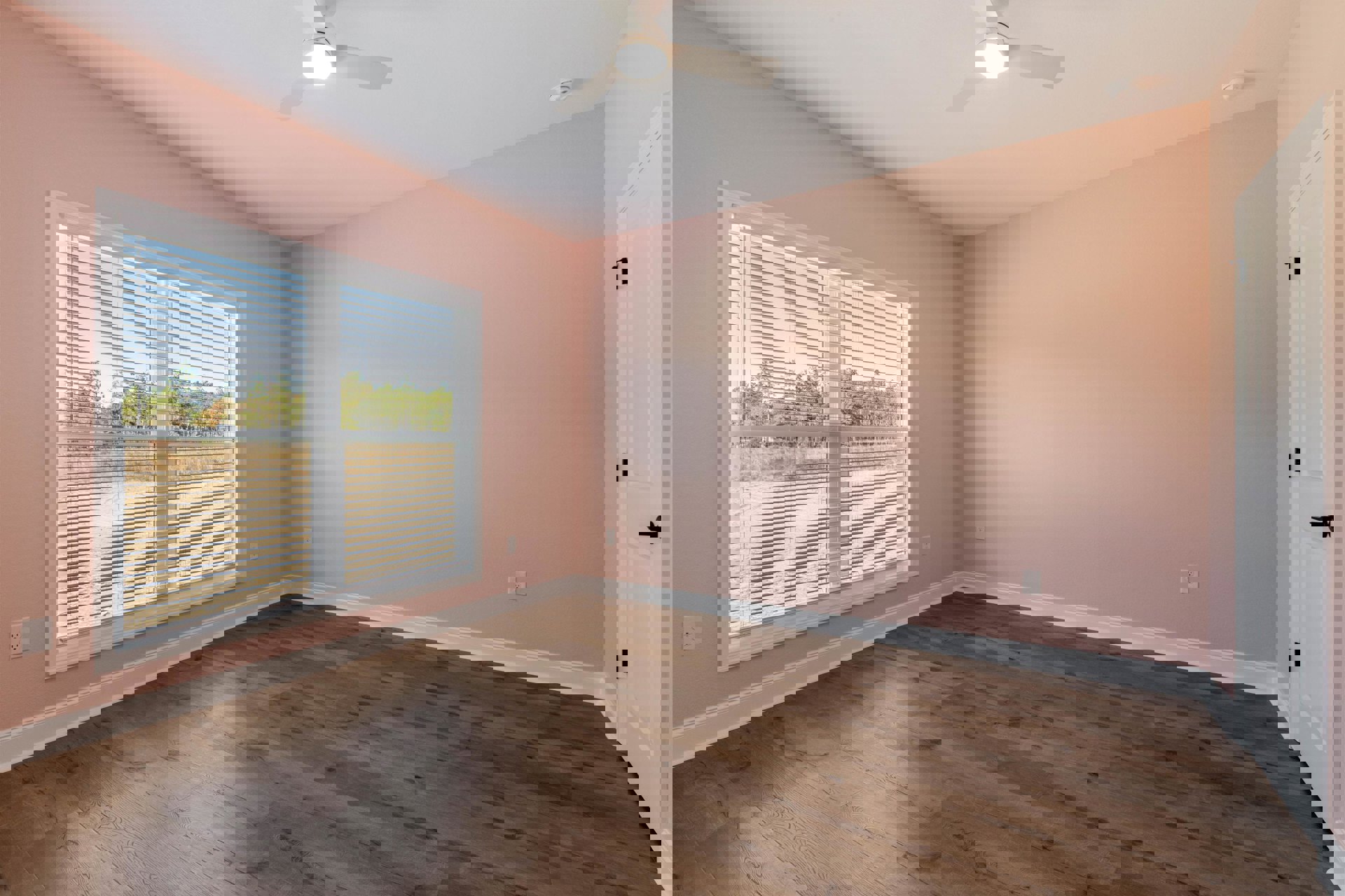 Bedroom with pink plaster walls, wood laminate flooring, window with white blinds, ceiling fan with light fixture, and white door featuring a black handle