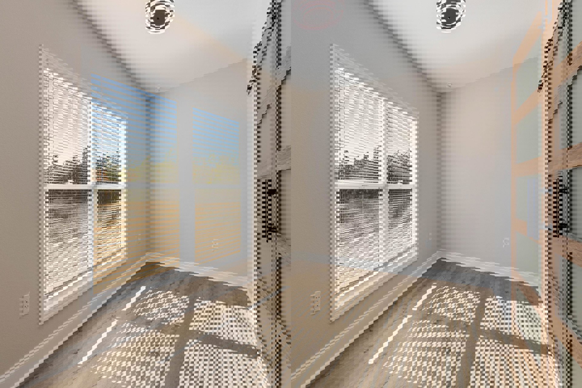 Sunlit room with white walls, wooden floor, large window fitted with horizontal blinds, modern light fixture, electrical outlet, and brushed metal door handle