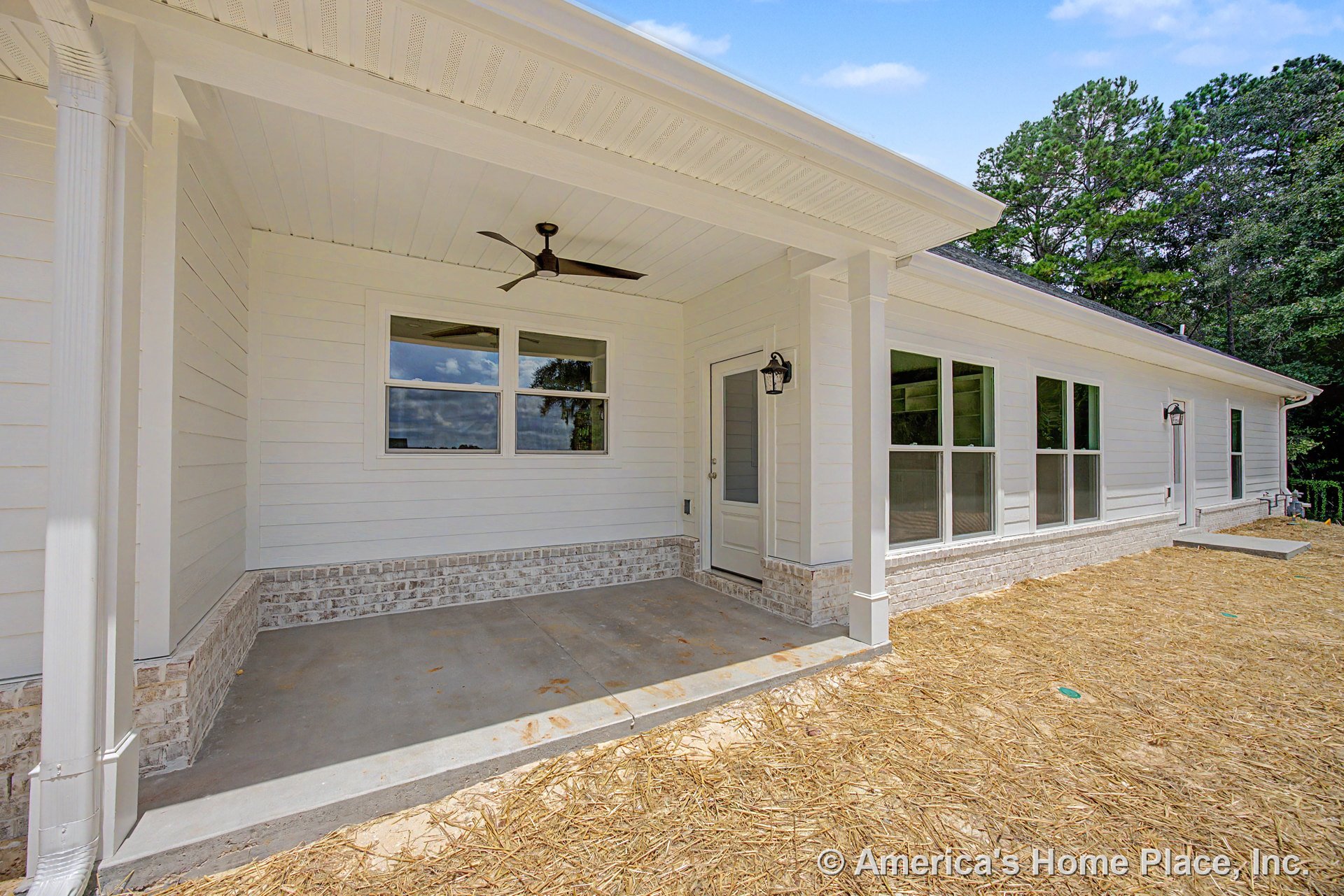 Covered front porch with white horizontal siding, brick foundation trim, large double windows, modern ceiling fan, glass exterior door, white columns, and concrete floor under