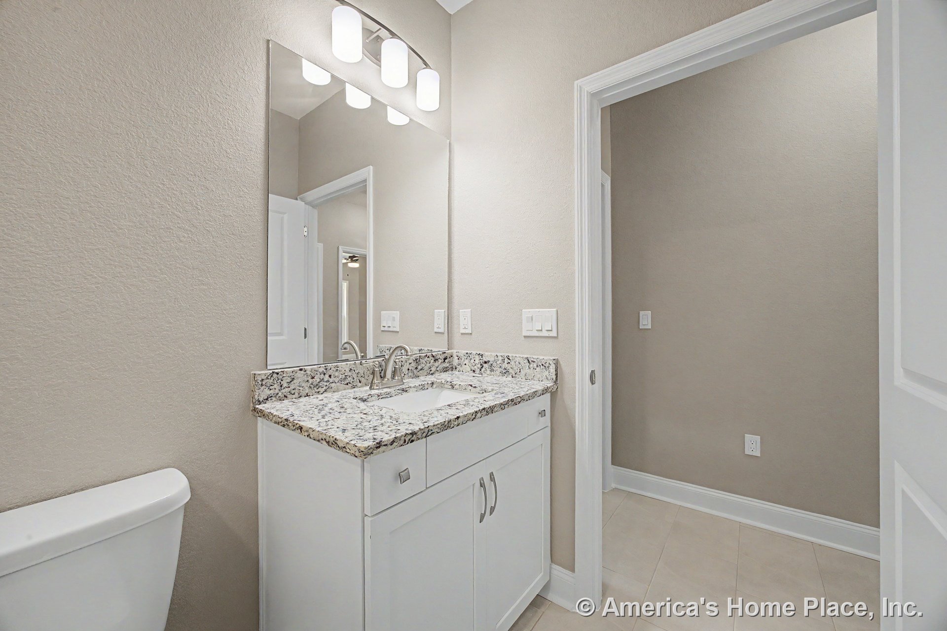 Granite countertop vanity with white cabinetry, rectangular wall mirror, built-in overhead lighting, neutral tile flooring, and trimmed doorways.