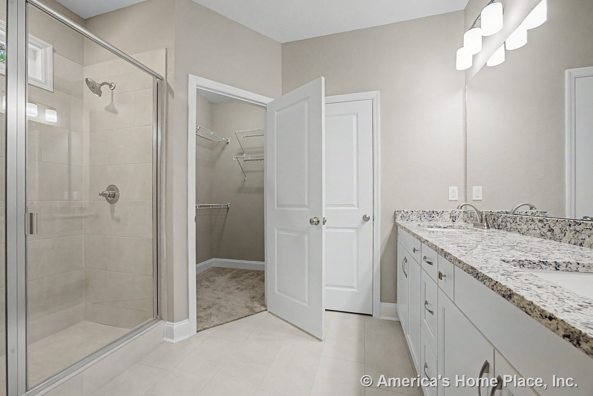 Glass-enclosed shower beside dual-sink granite vanity with white cabinetry, modern overhead lighting, tile flooring, and walk-in closet featuring wire shelving and interior doors.