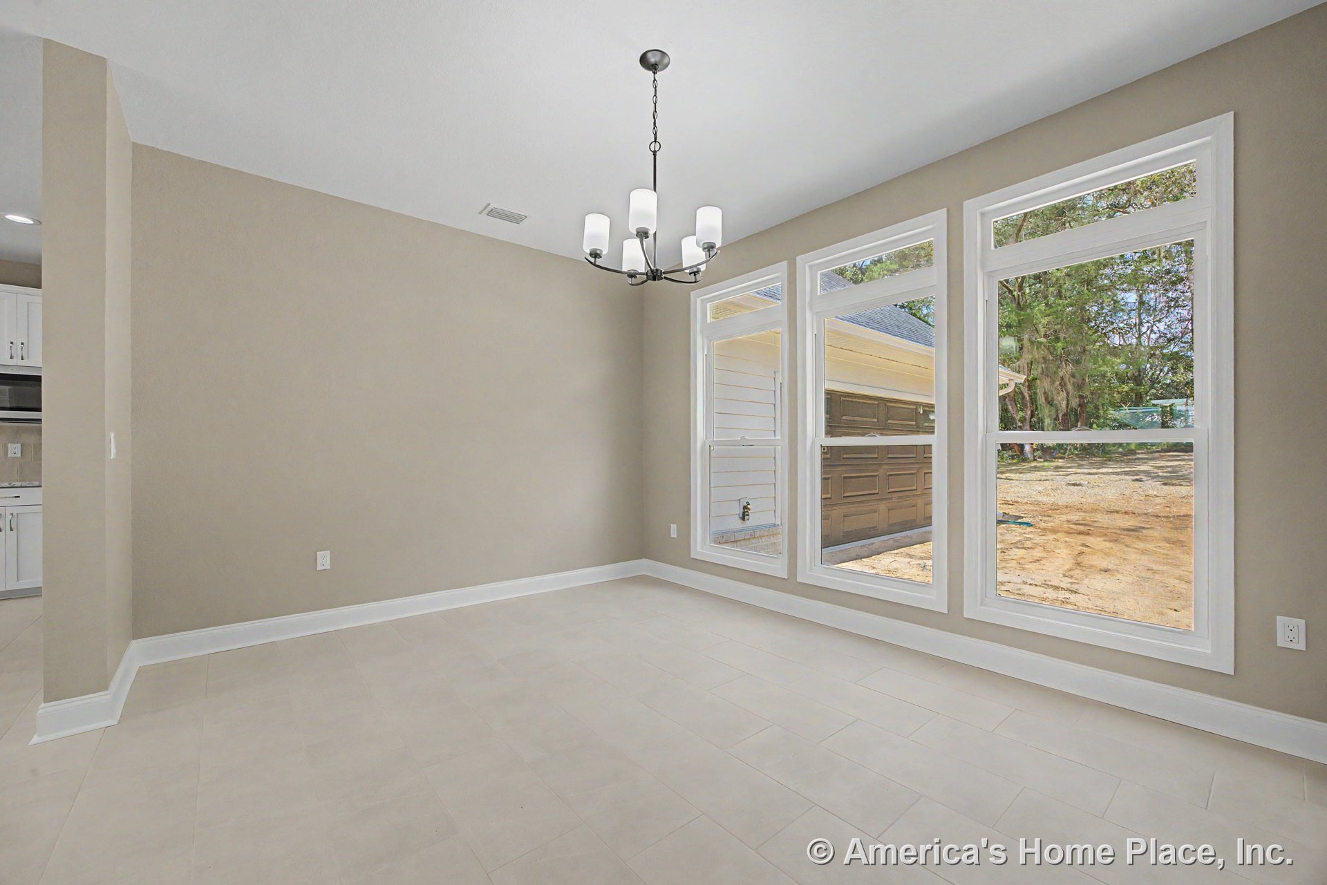 Dining area with three tall picture windows, neutral tile flooring, modern chandelier, white window trim, beige walls, baseboard trim, and adjacent kitchen cabinetry in an open