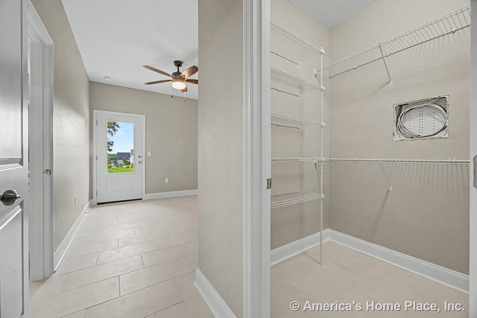 Hallway with light tile flooring and white baseboards leads to a walk-in closet featuring built-in wire shelving; glass exterior door and ceiling fan with light fixture; neutral