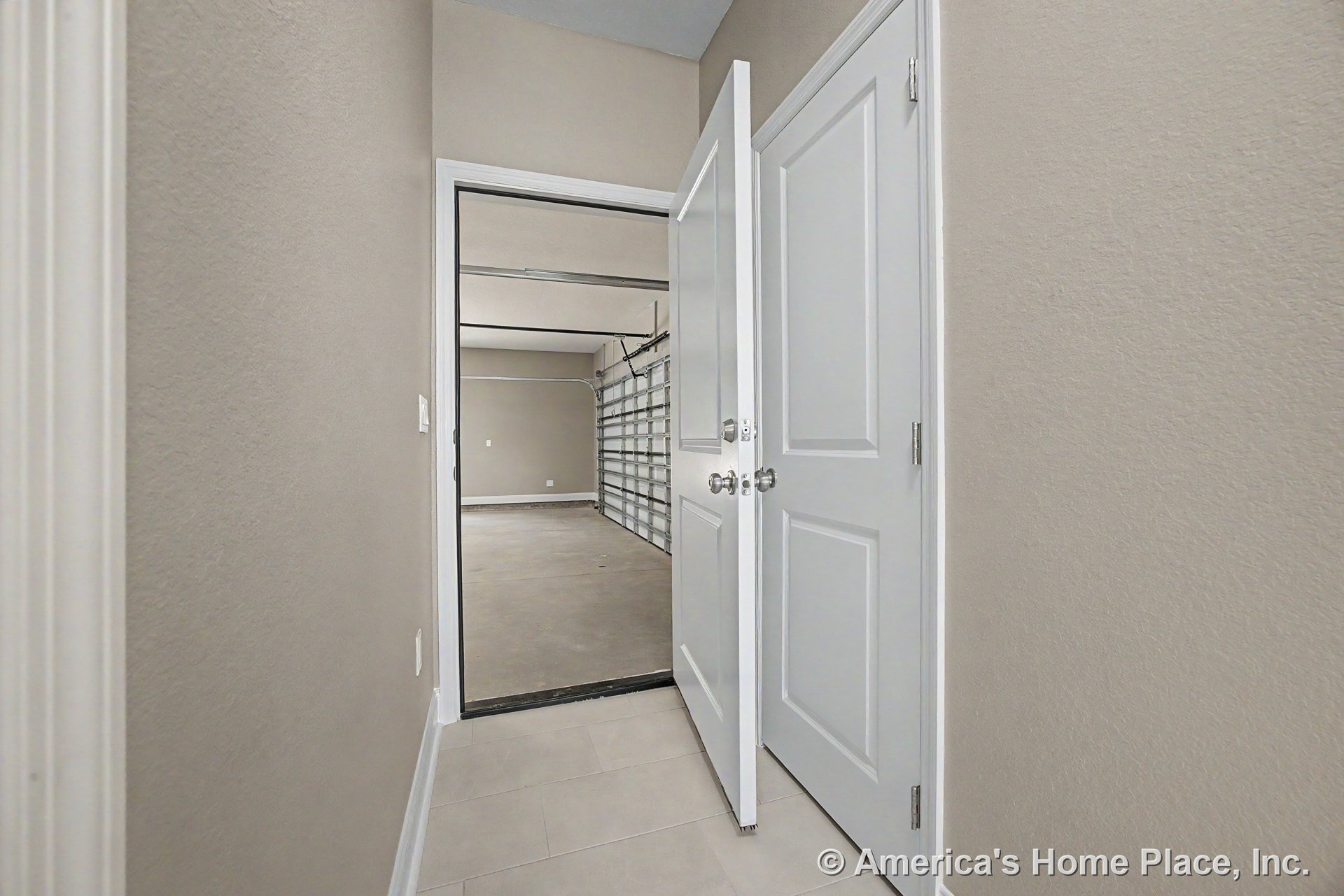 Hallway with beige painted walls, white baseboard trim, tile flooring, and a raised ceiling, leading to a garage through a white paneled interior door; garage features a concrete