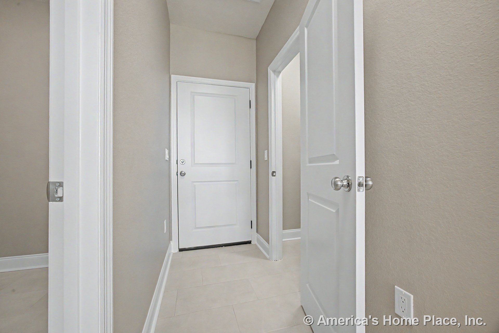 Hallway with beige tile flooring, light gray walls, white paneled doors featuring brushed metal knobs, and white baseboards.