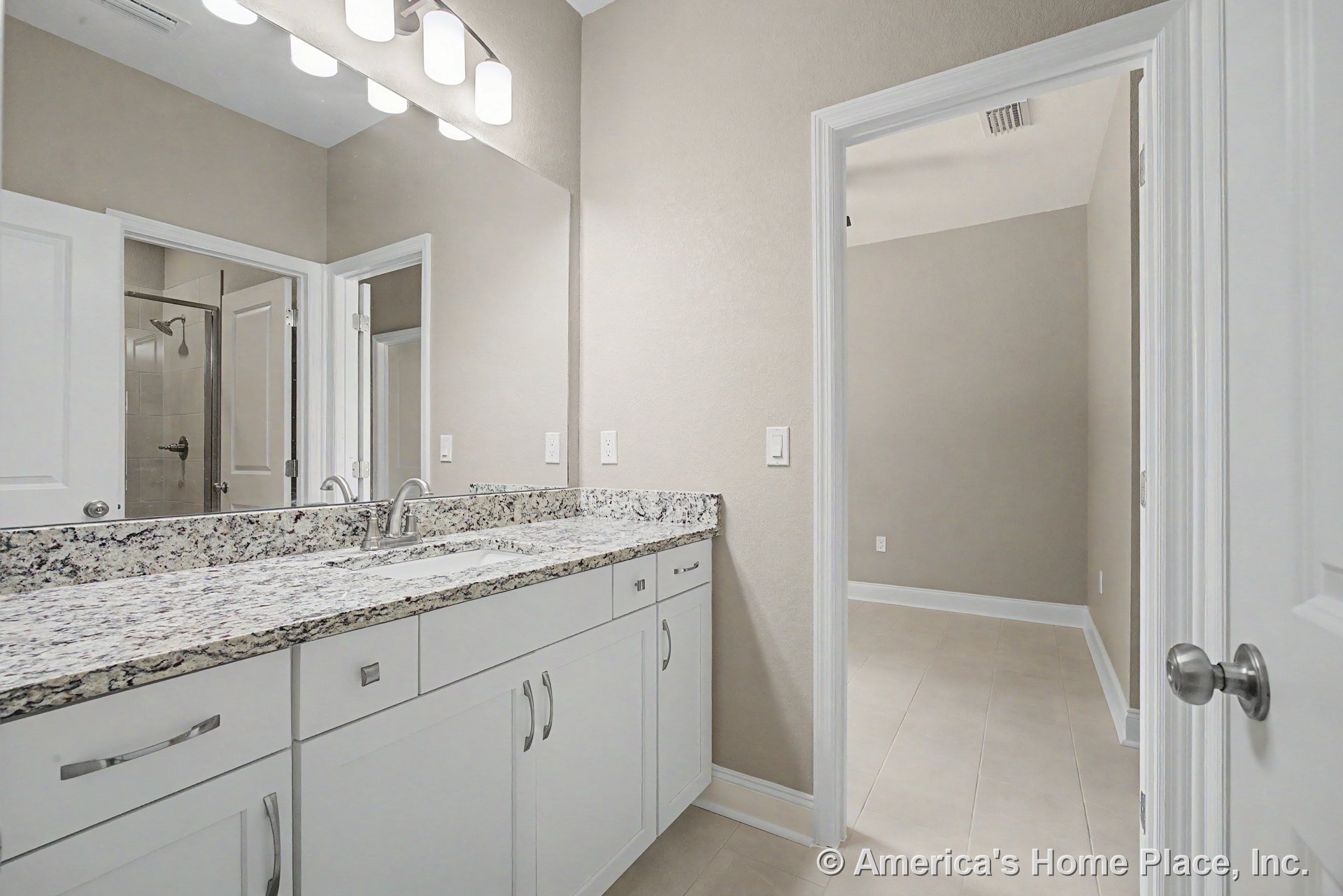 Granite double sink vanity with white shaker cabinets, expansive wall mirror, built-in lighting above, neutral tile flooring, and modern door hardware.