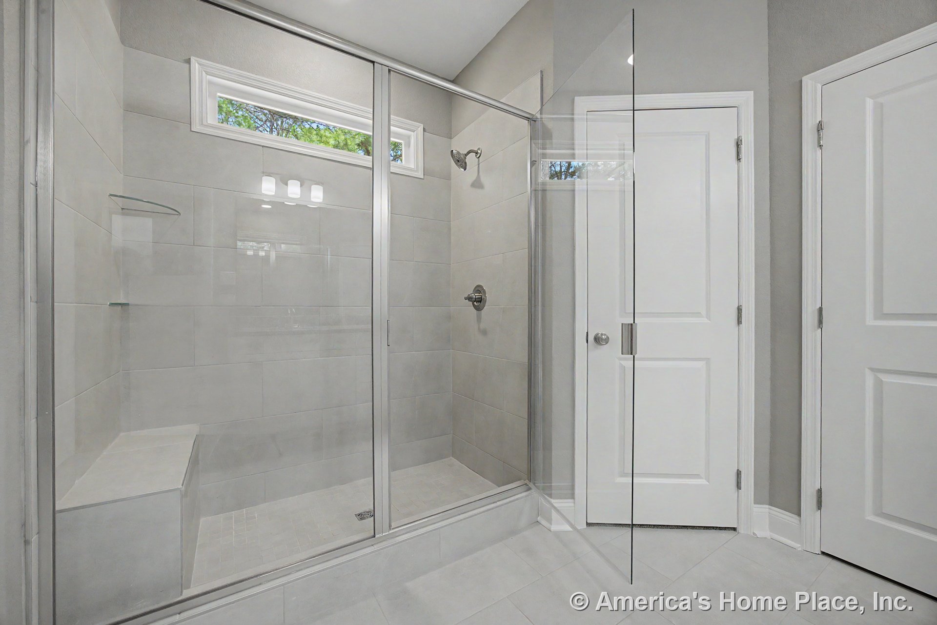 Spacious bathroom with walk-in shower featuring glass enclosure, built-in bench, light gray tile walls, transom window, and white paneled doors.